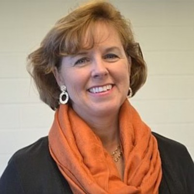 A woman with short brunette hair, a black top, and orange scarf smiles at the camera