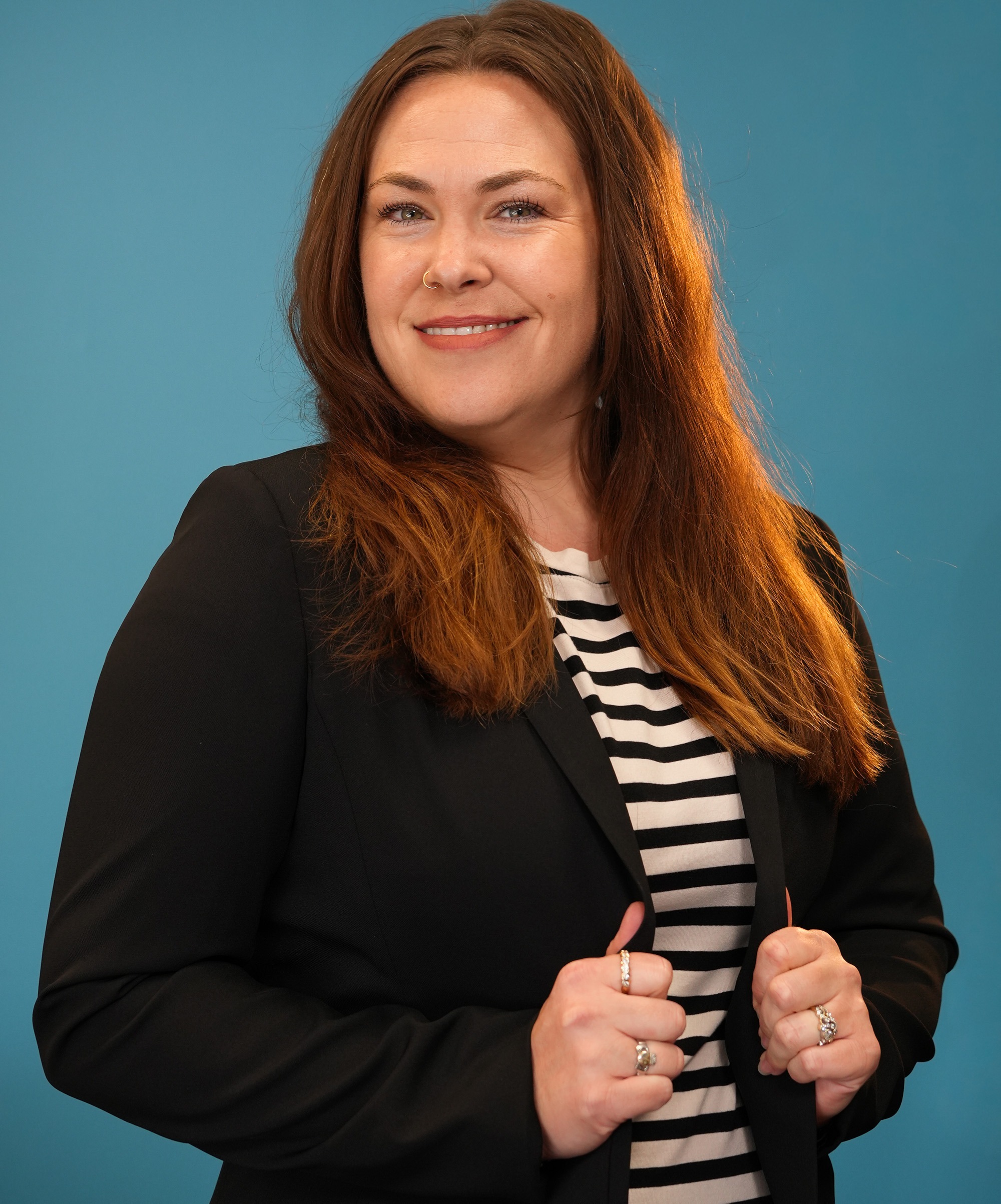 A brunette woman with a black and white striped shirt and a black blazer smiles at the camera, with a teal backdrop.