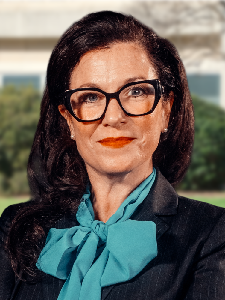 A woman with dark brown hair and black glasses, stands smiling wearing a black blazer with a teak scarf