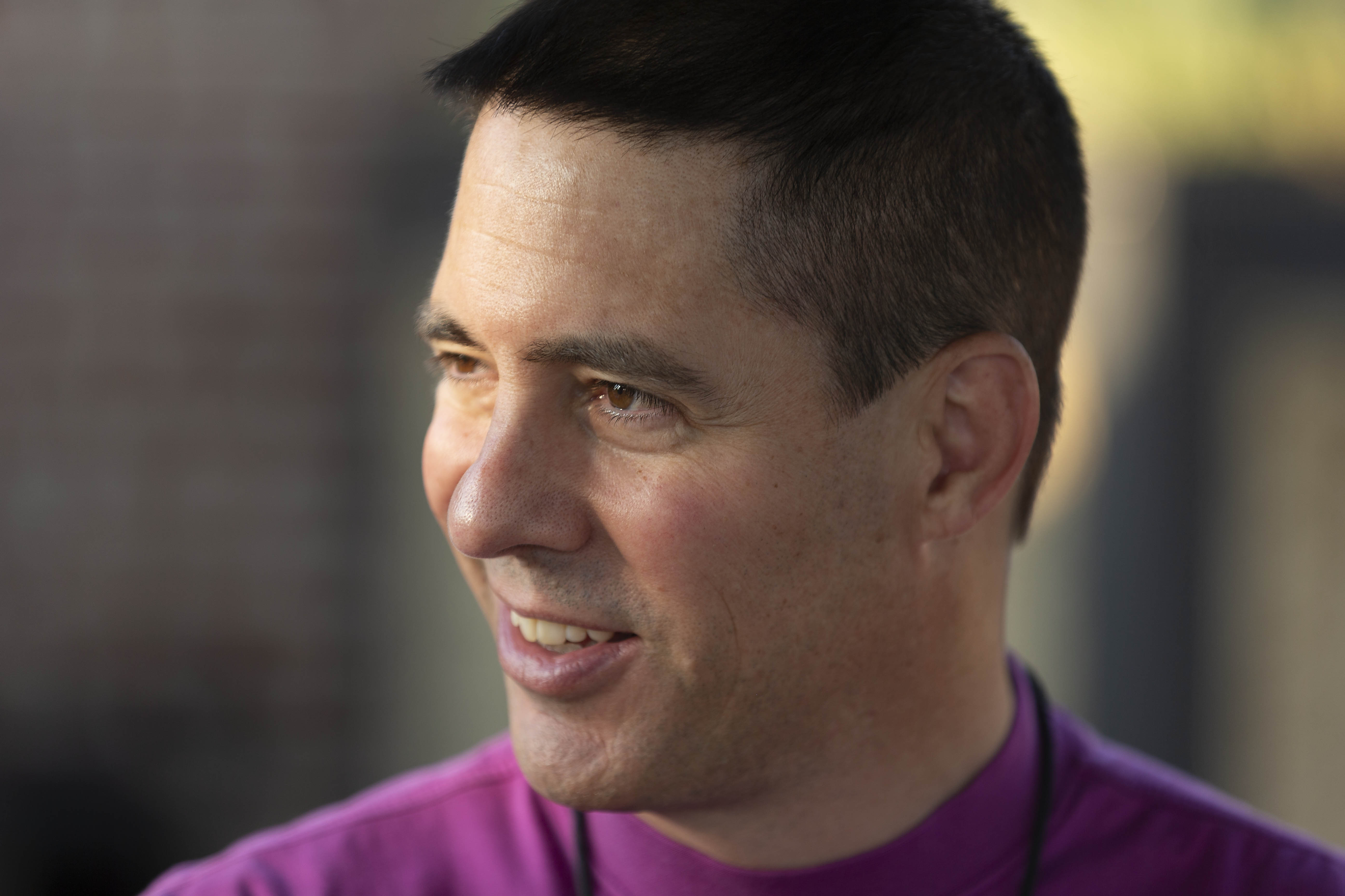A man with dark hair and a pink shirt looks to the right with a smile.