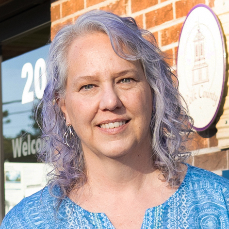 A woman with lavender hair and a blue shirt stands smiling in front of a brick building.