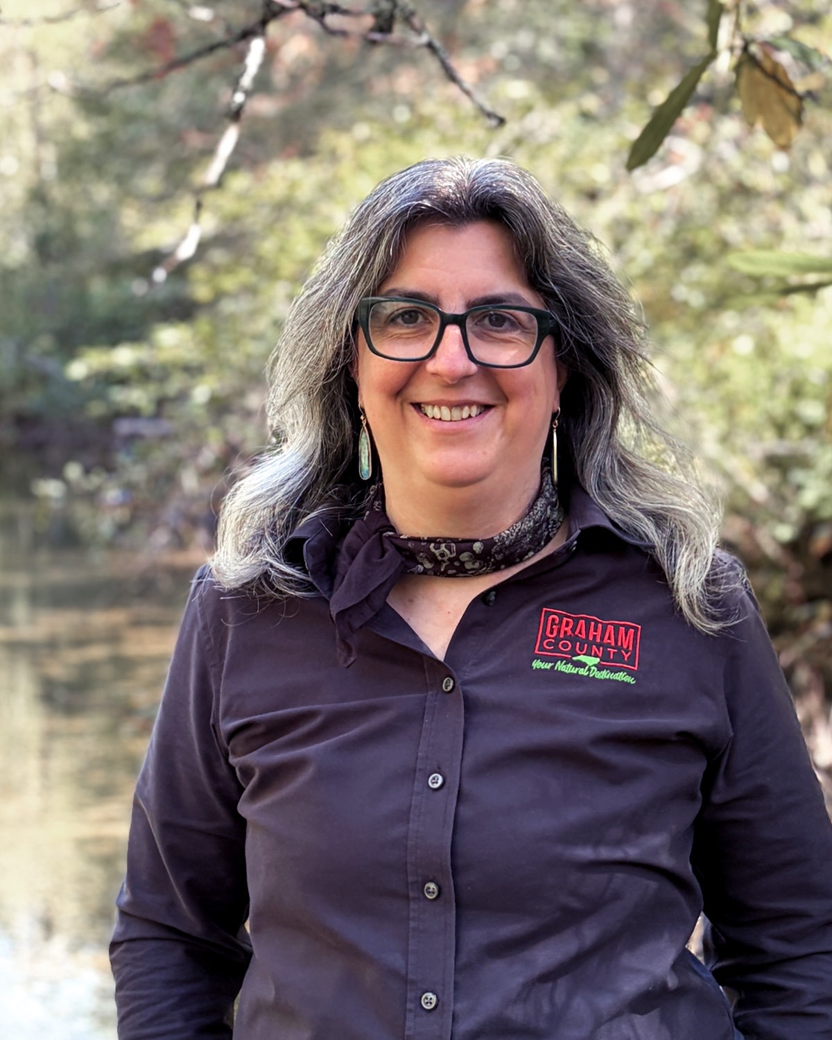 A woman with dark gray hair and glasses stands in a forest setting wearing a black shirt, smiling at the camera