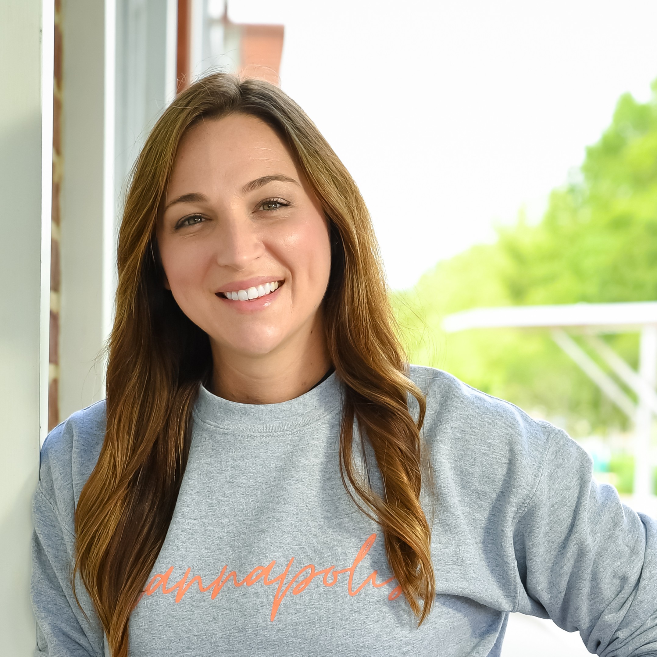 A woman with brown hair and a gray sweatshirt leans against a brick building and smiles at the camera.