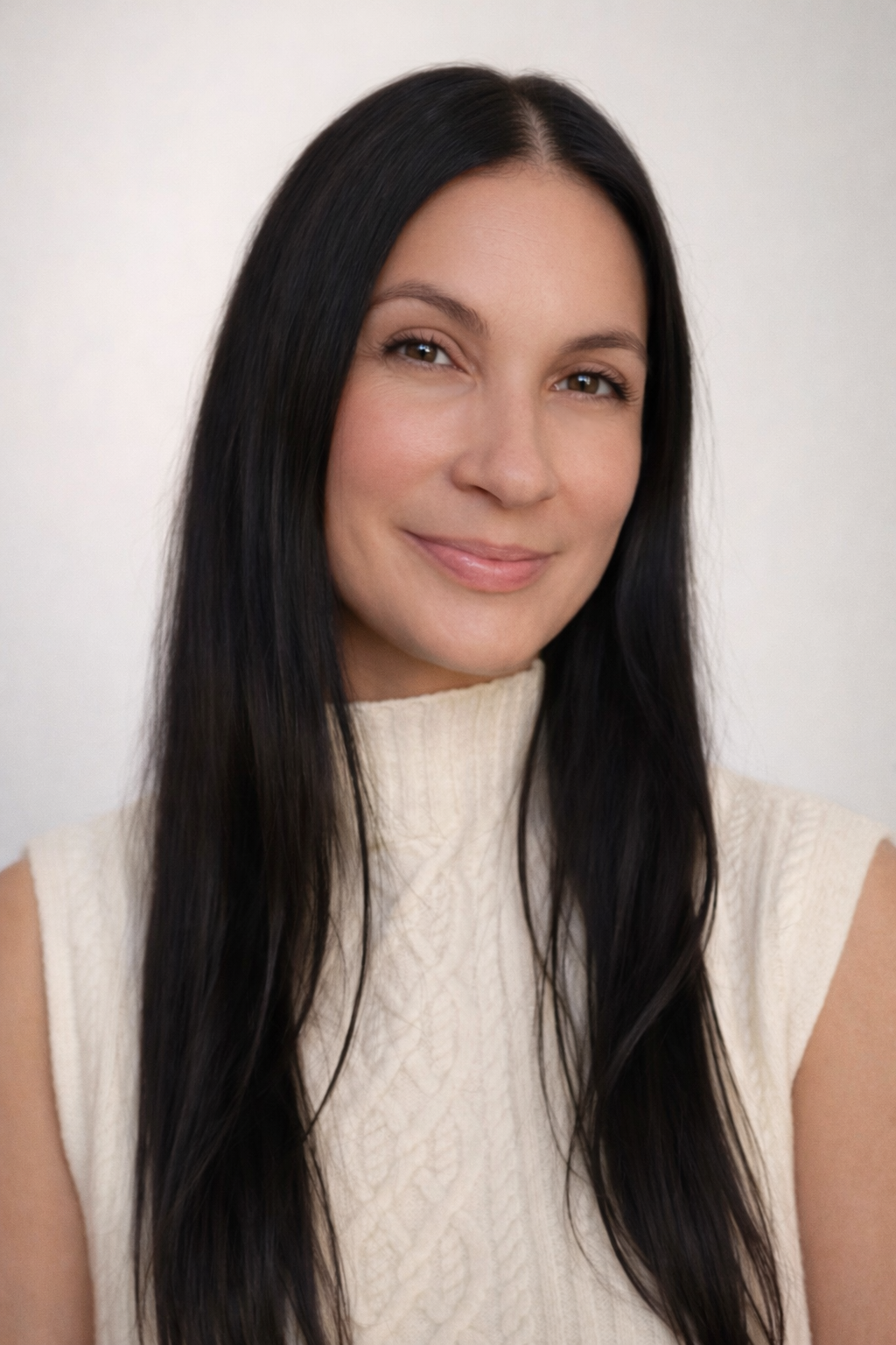 A woman with long black hair and a cream sleeveless sweater smiles at the camera