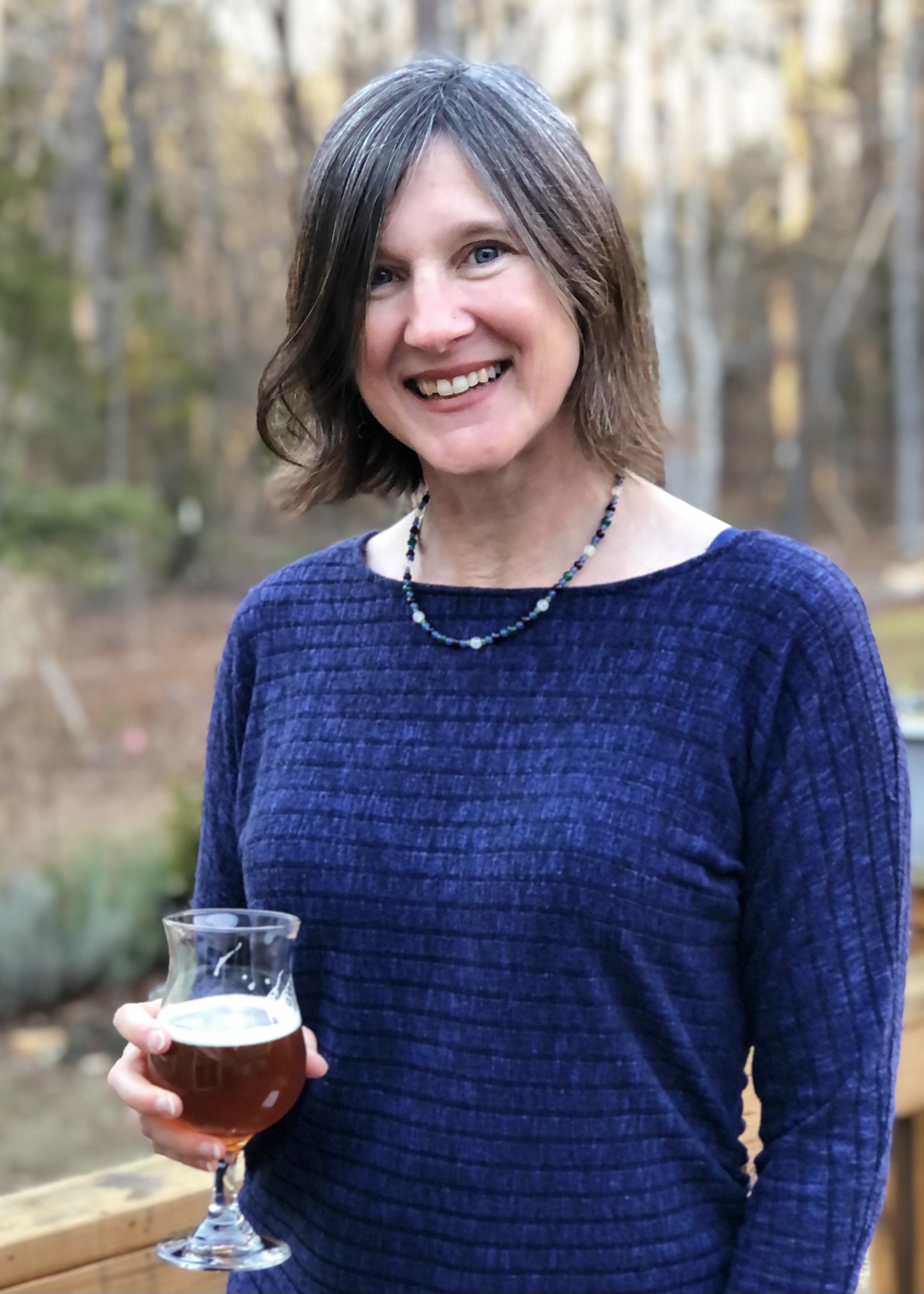 A woman with short gray hair and a blue shirt stands smiling with glass on beer in her hand