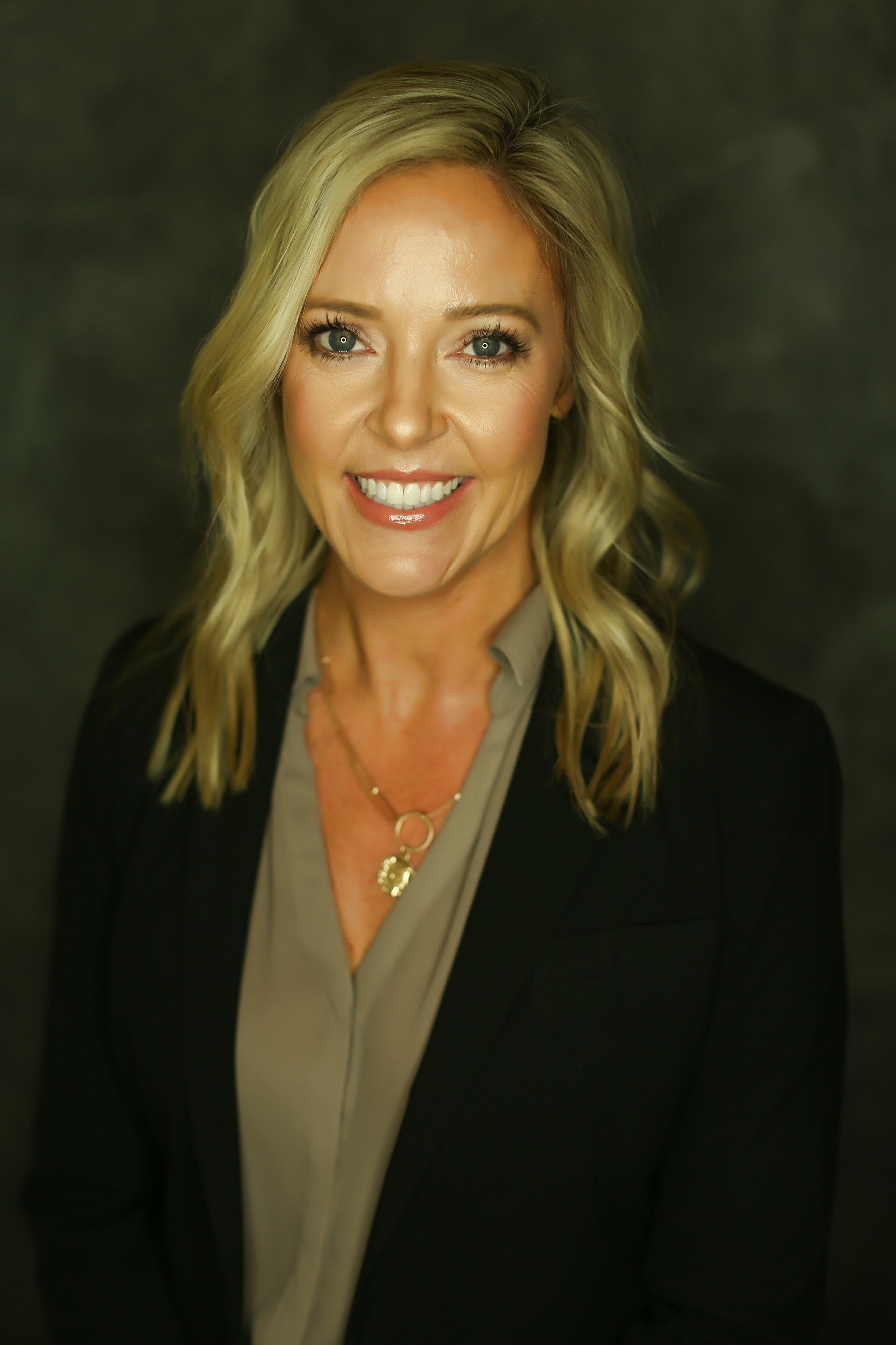 A blonde woman with a silver shirt and a black blazer smiles up at the camera