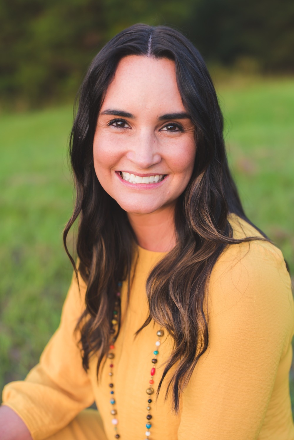 A woman with brown hair and a yellow shirt smiles at the camera with grass in the background
