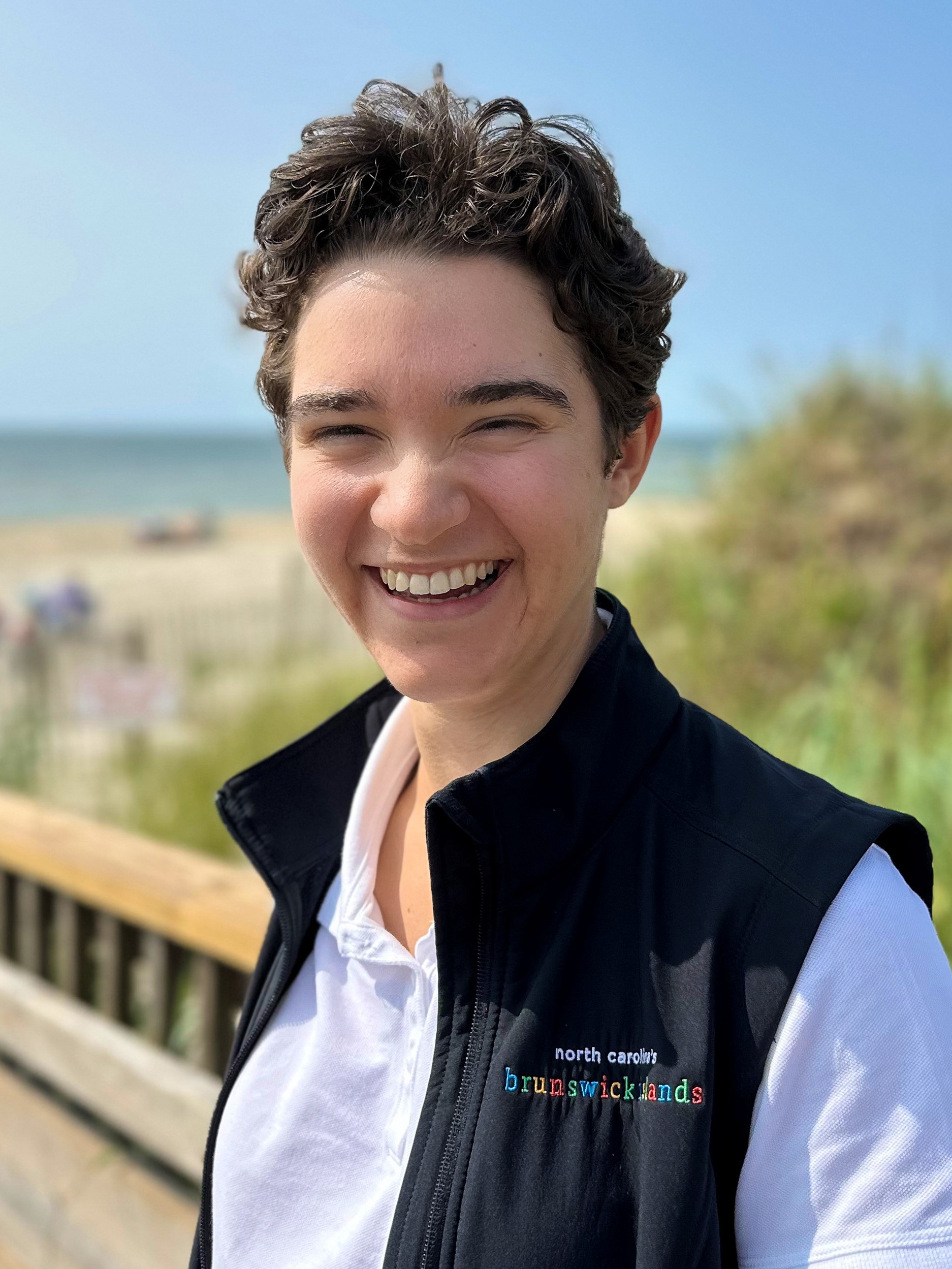 A woman with short brown hair wearing a white shirt with a black vest overtop, smiles at the camera with a beach and ocean in the background
