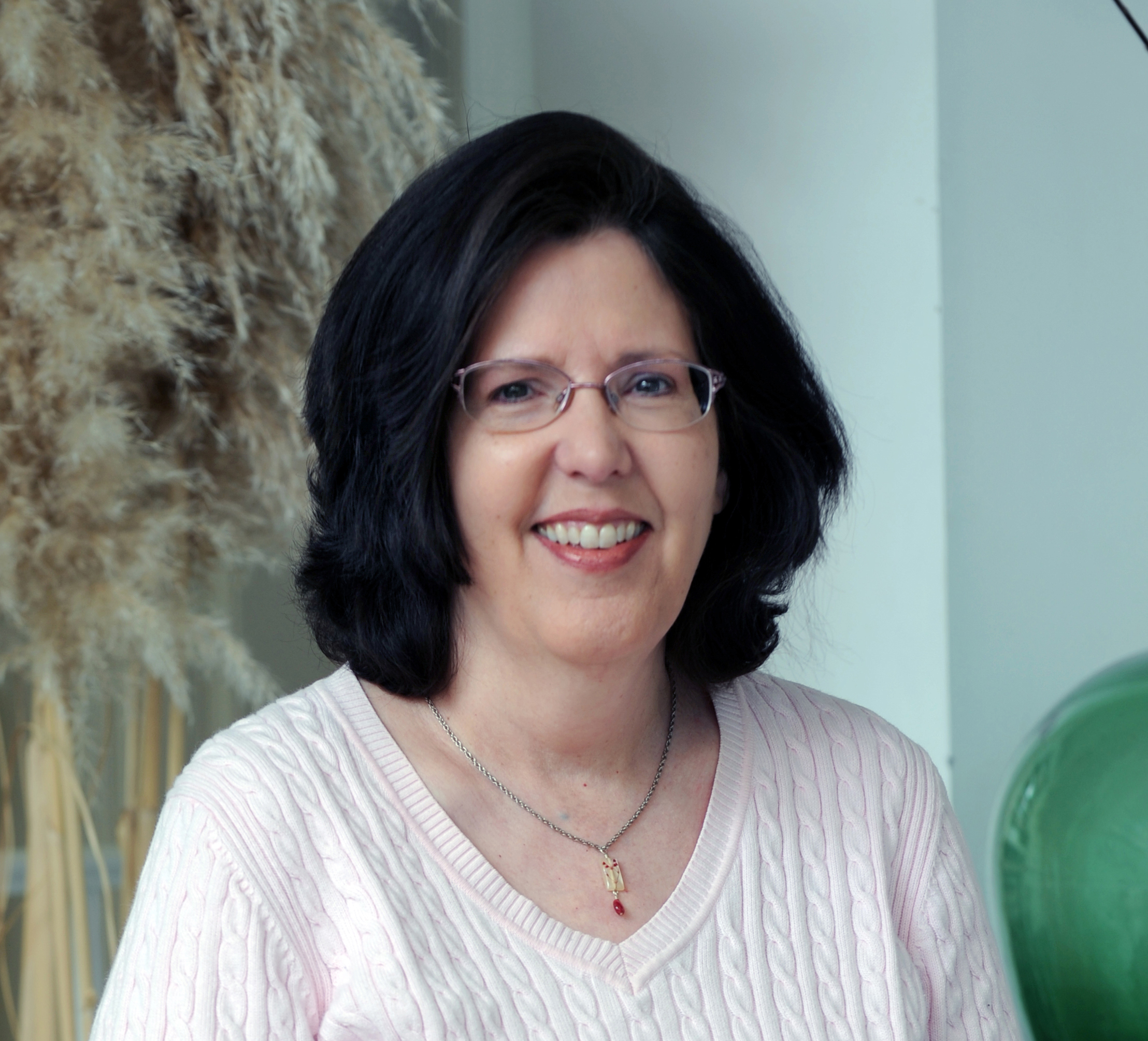 A woman with black hair and glasses, wearing a light pink sweater, smiles at the camera.