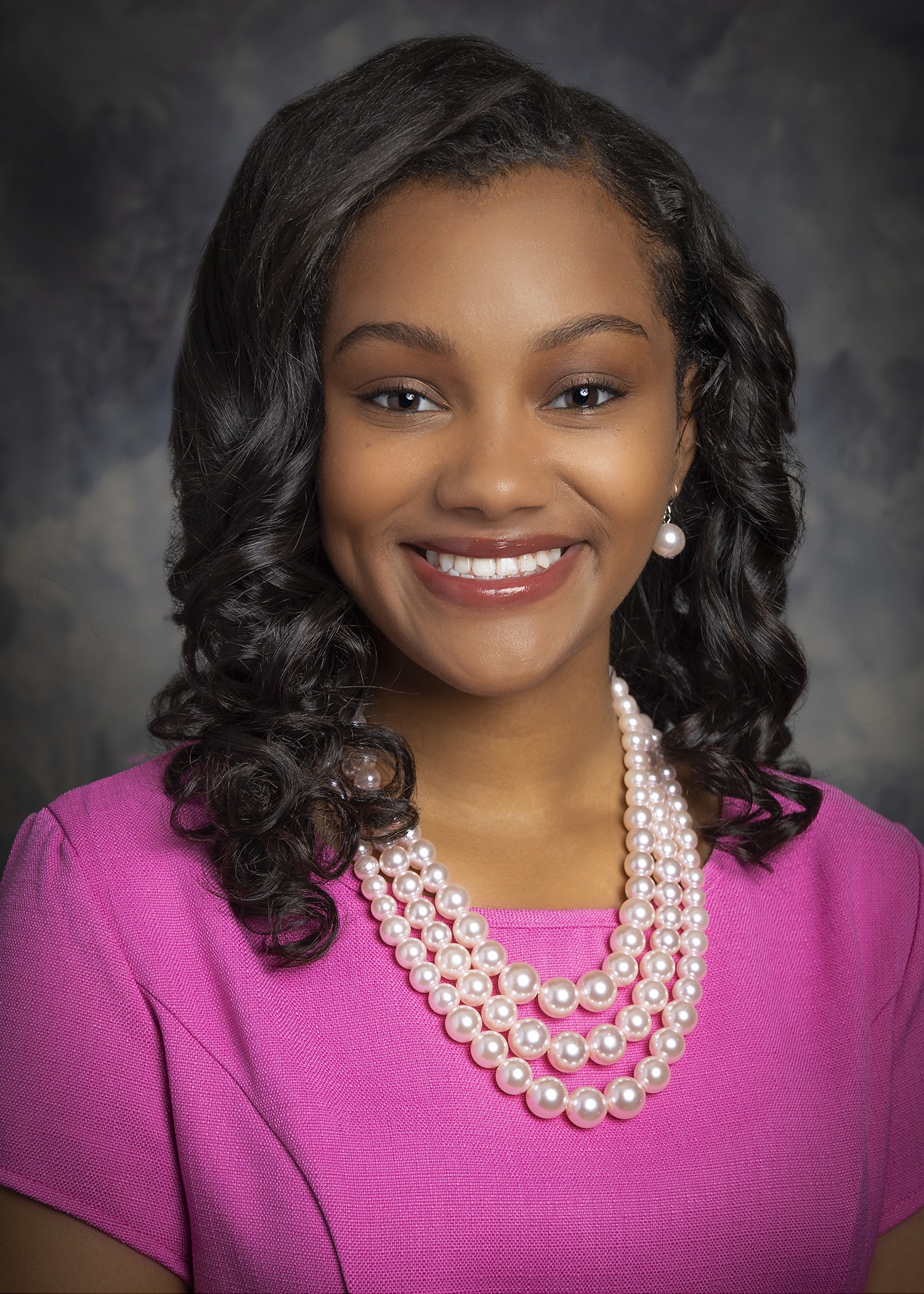 A woman with black hair, a large pearl necklace and a bright pink shirt, smiles at the camera.