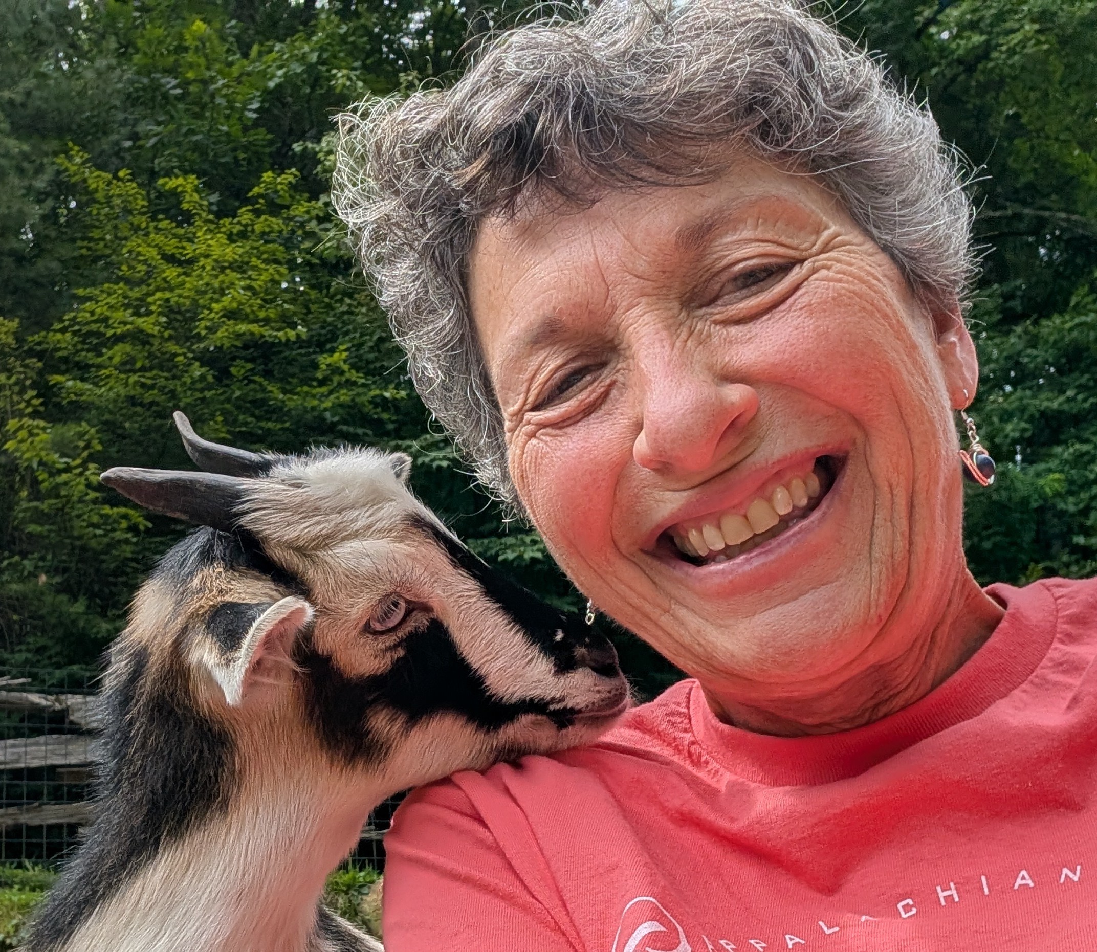 A woman with short gray hair and a pink shirt smiles at the camera, while a small goat rests it's chin on her shoulder.