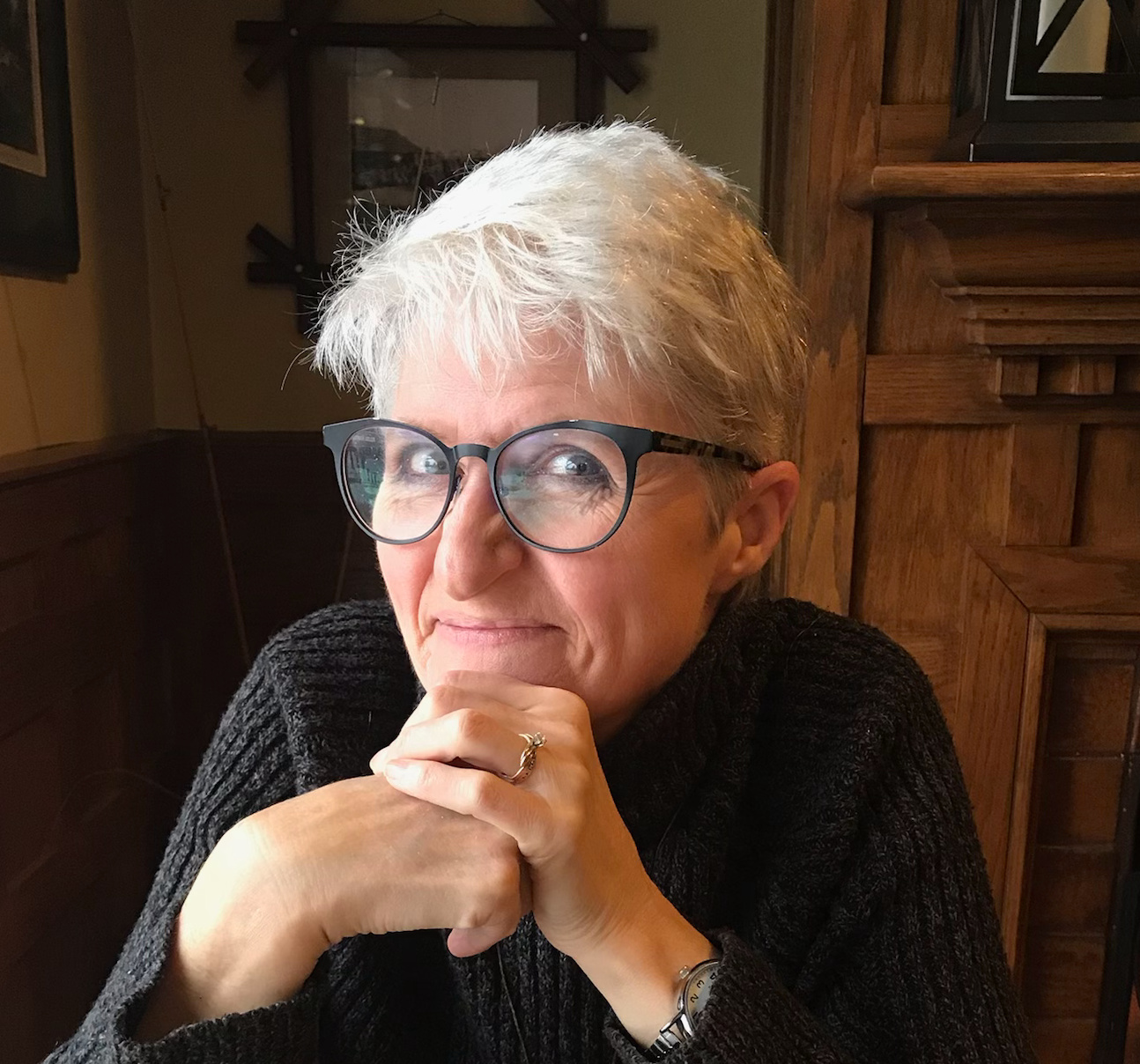 A woman with short gray hair in a black sweater with back glasses, smiles at the camera