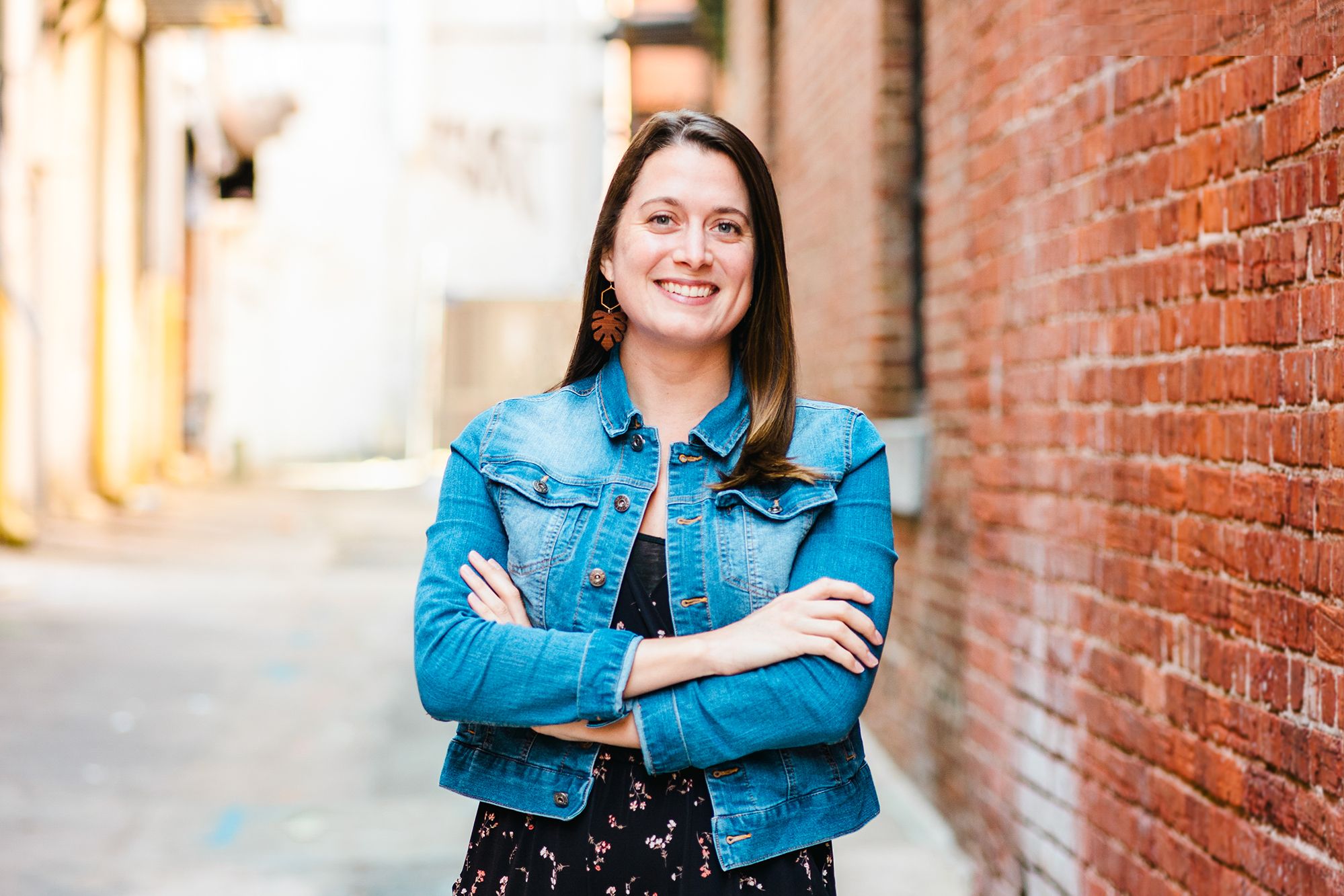 A brunette woman with a jean jacket and a black floral dress, stands with her arms crossed behind a brick building and smiles at the camera.