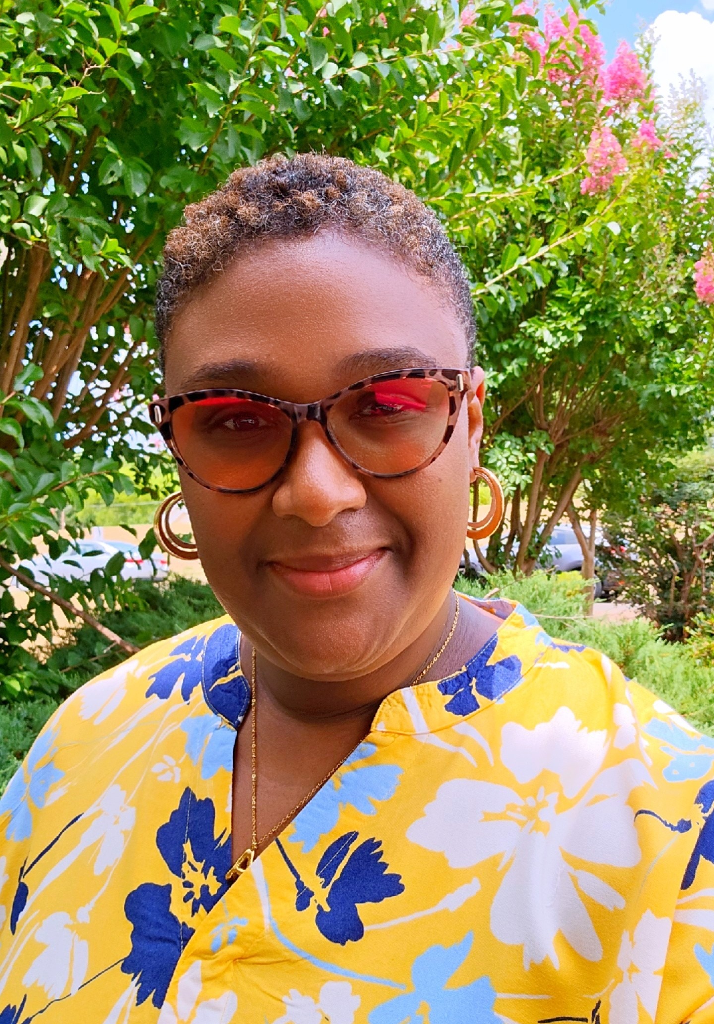 A woman with orange-tinted sunglasses and a yellow and blue floral shirt stands in front of foliage while smiling