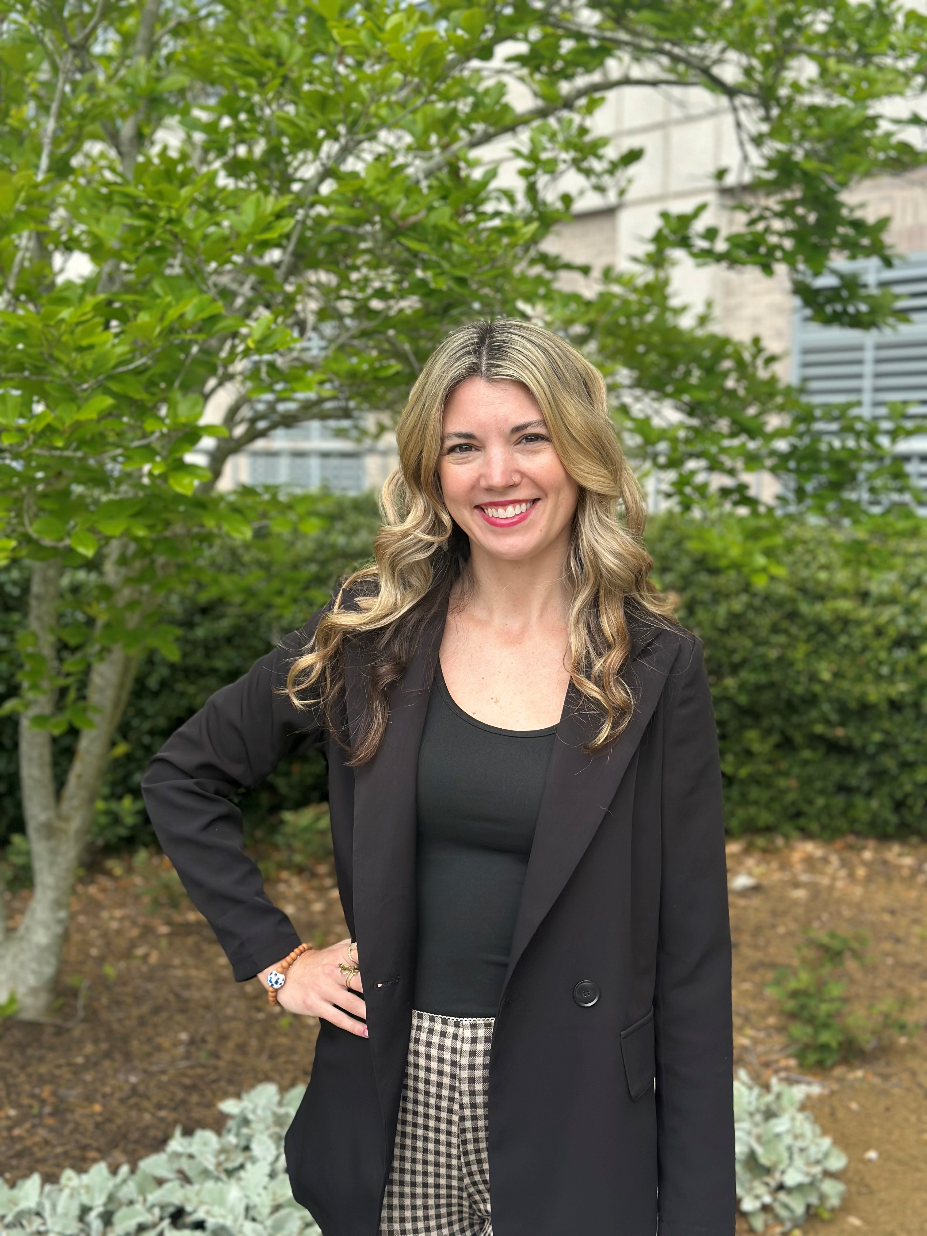A woman with brunette hair wearing a black blazer and pants stands with one hand on her hip in front of bushes and smiles