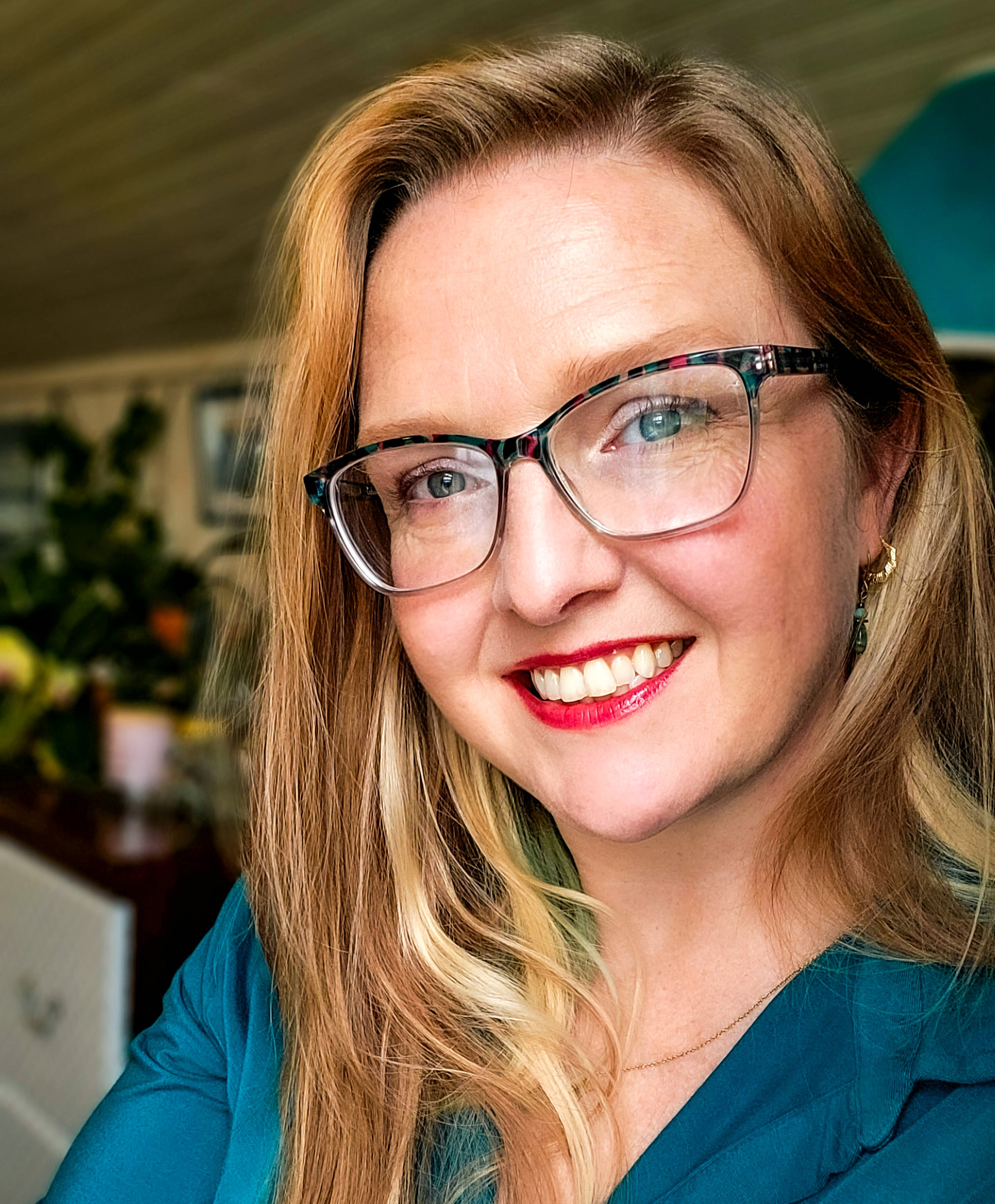 A blonde woman with red lipstick and a teal shirt smiles at the camera