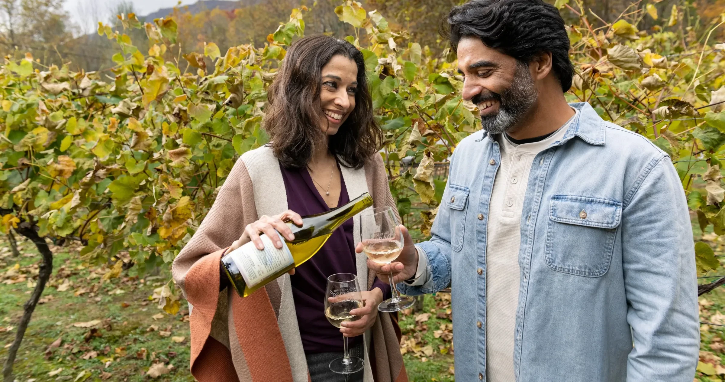 Woman smiling while pouring white wine into her partner's glass within row of grapes at Grandfather Vineyard & Winery.
