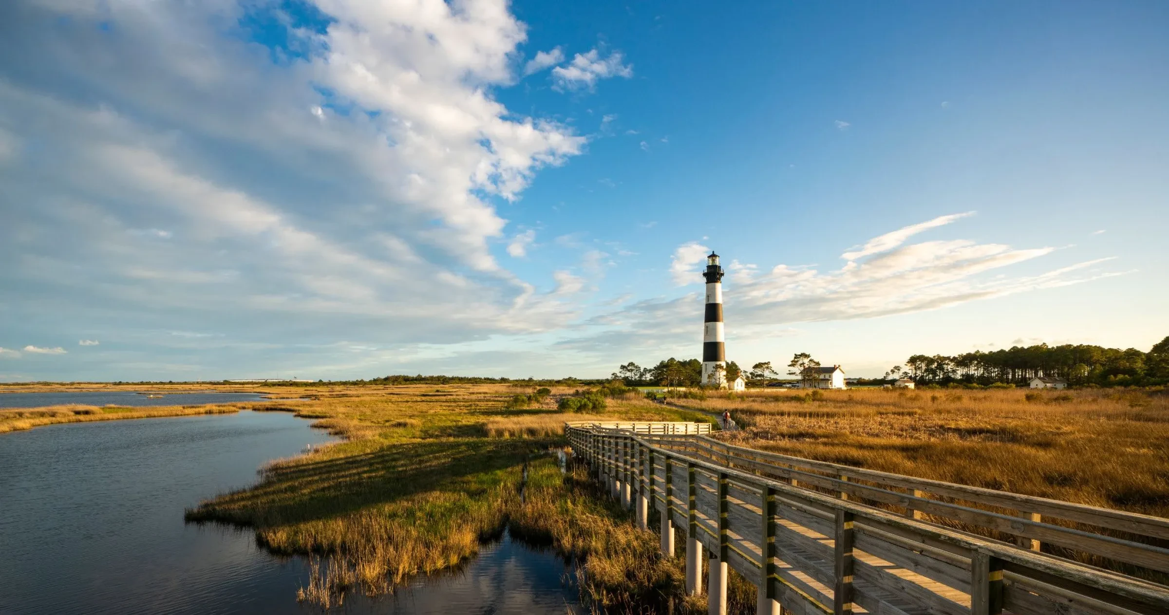 Golden hour view of Bodie Island Lighthouse with pier and sound. Lighthouse placed slightly right of center frame.