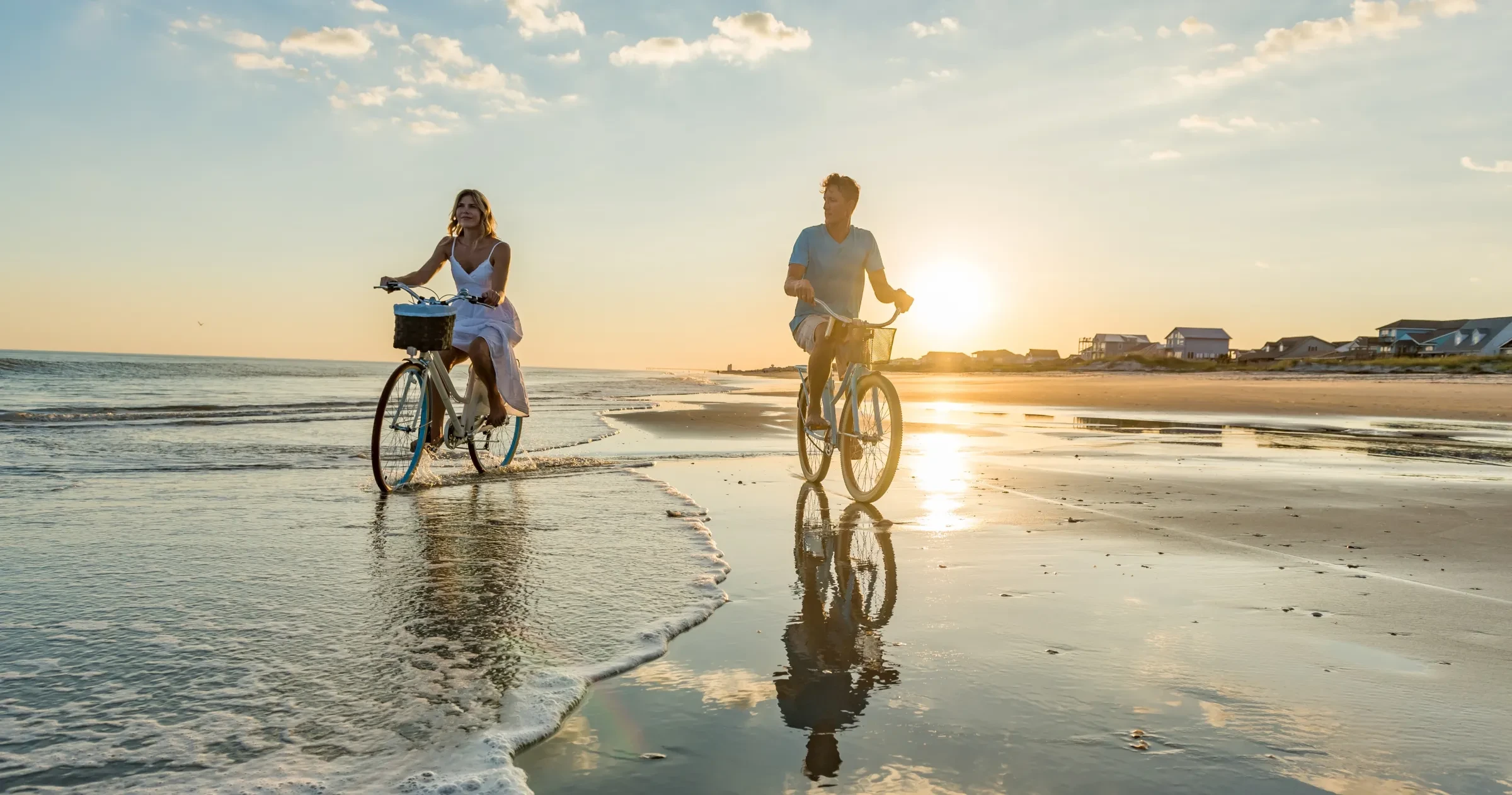 Couple riding bikes on beach in Ocean Isle Beach as sun sets in background.