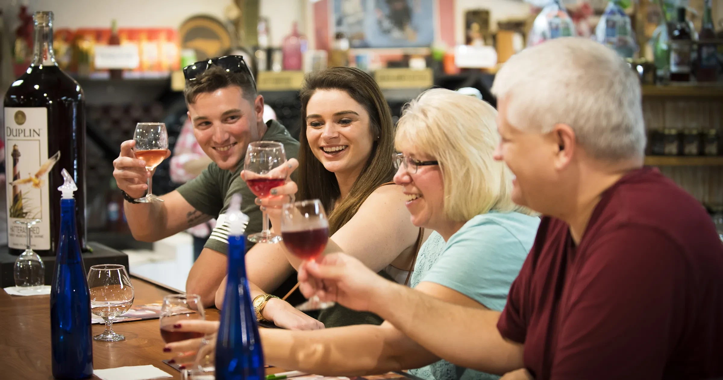 A group of four people sit at a bar, smiling and raising glasses of red wine in a toast. Bottles and glasses are on the wooden counter in front of them, and shelves of wine are visible in the background.