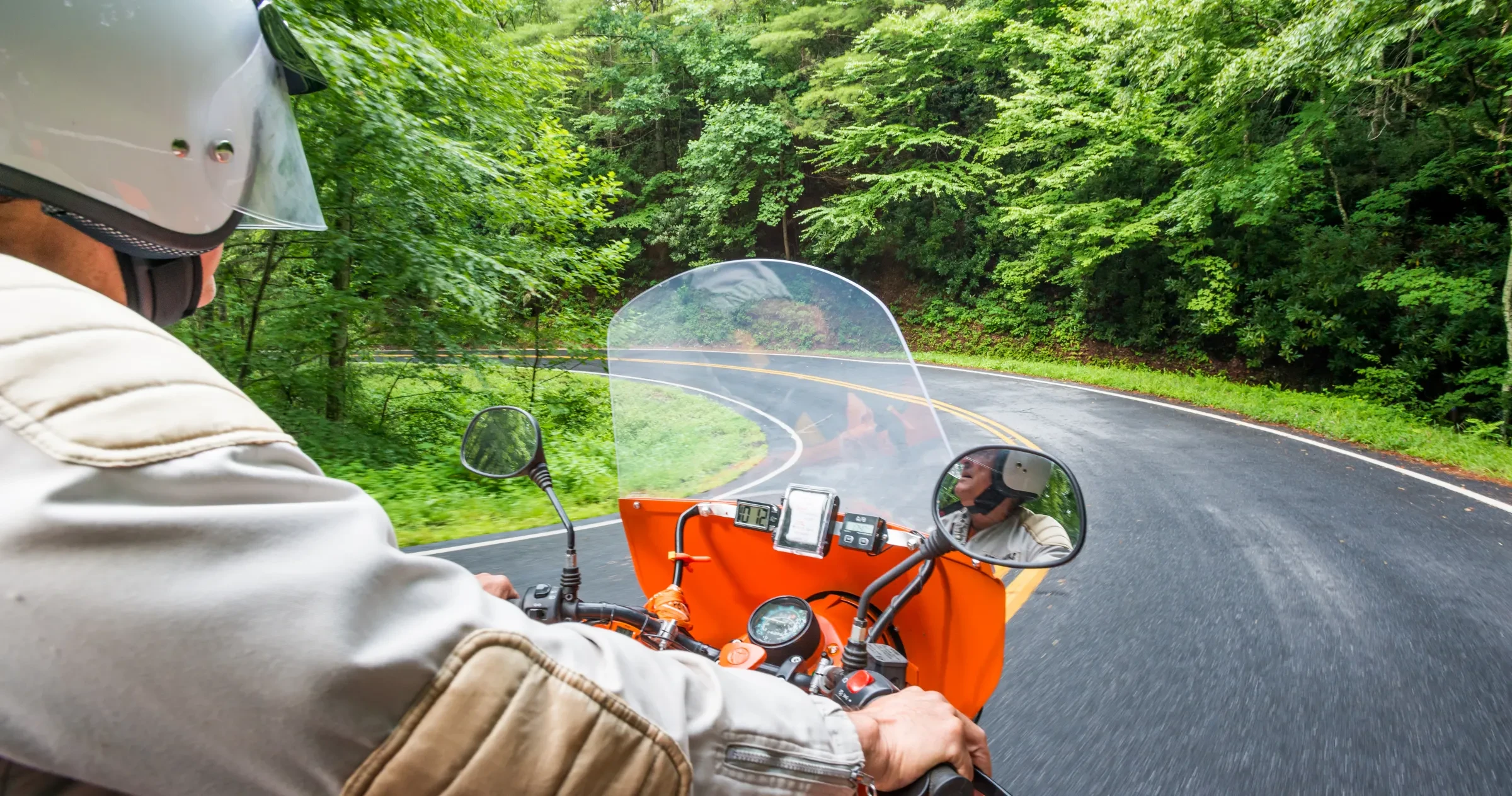 A person wearing a helmet rides an orange motorcycle with a sidecar along a winding road through lush green forest, seen from their perspective. The road curves ahead, and trees line both sides.