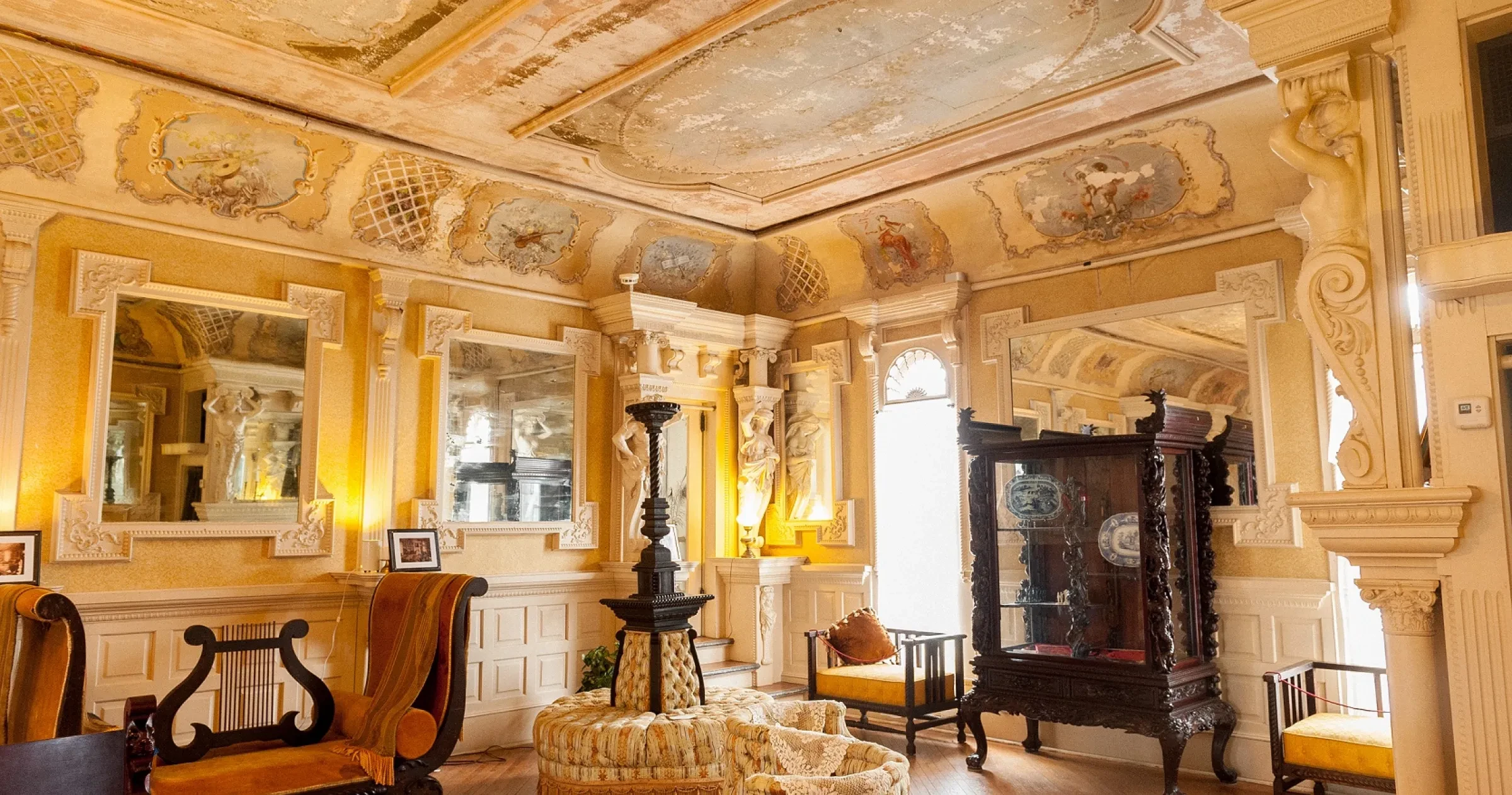 An ornate vintage room with painted ceilings, large mirrors, antique furniture, and elaborate wall decorations, filled with warm natural and artificial light.