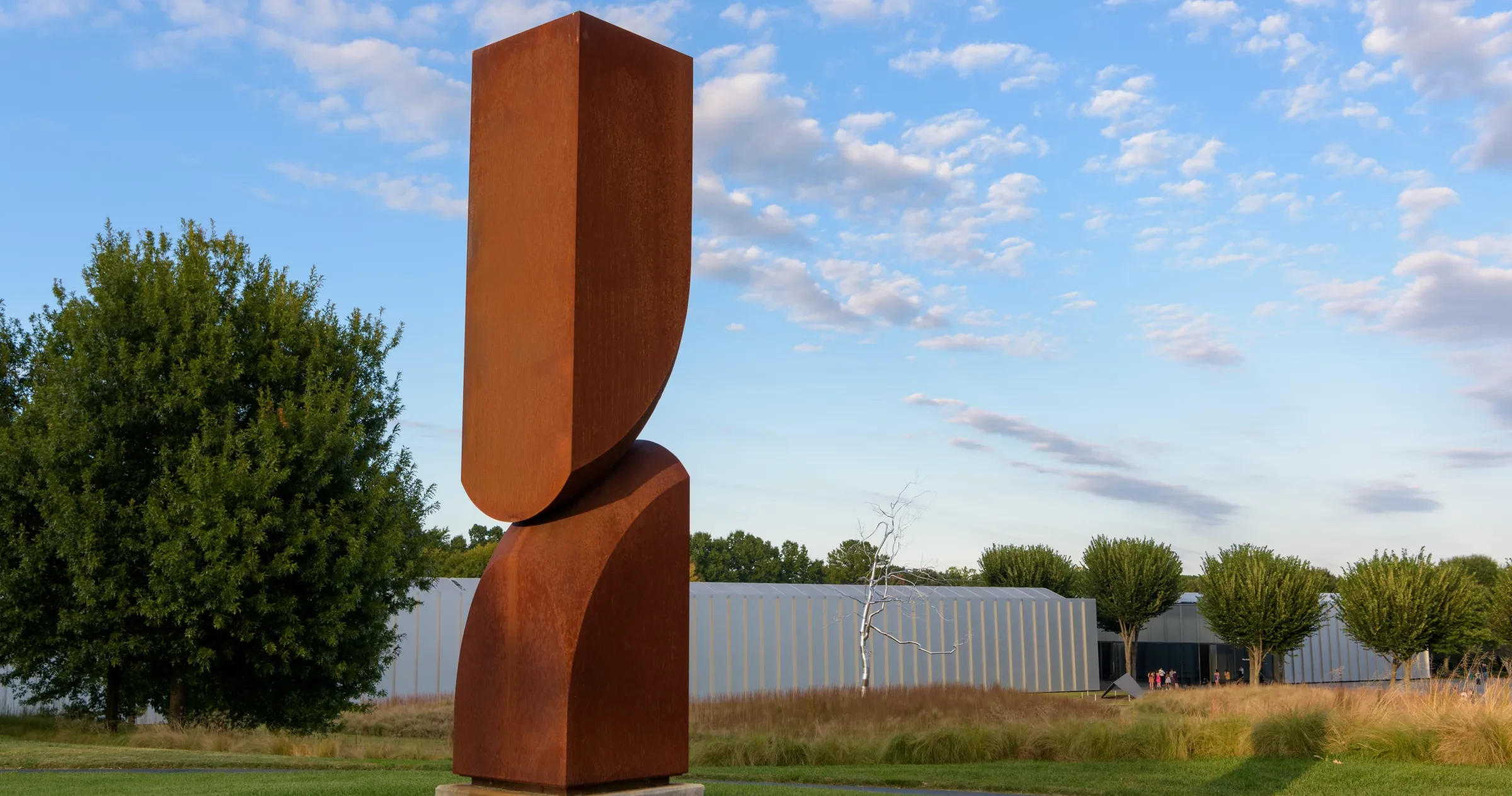 A tall, rust-colored abstract metal sculpture stands on a grassy lawn with trees and a modern building in the background under a partly cloudy sky.