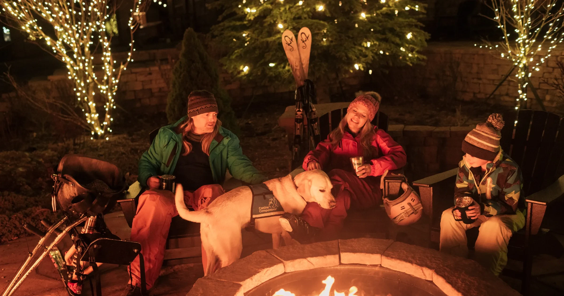 Family and dog sitting around ski resort fire at night surrounded by lit-up trees.