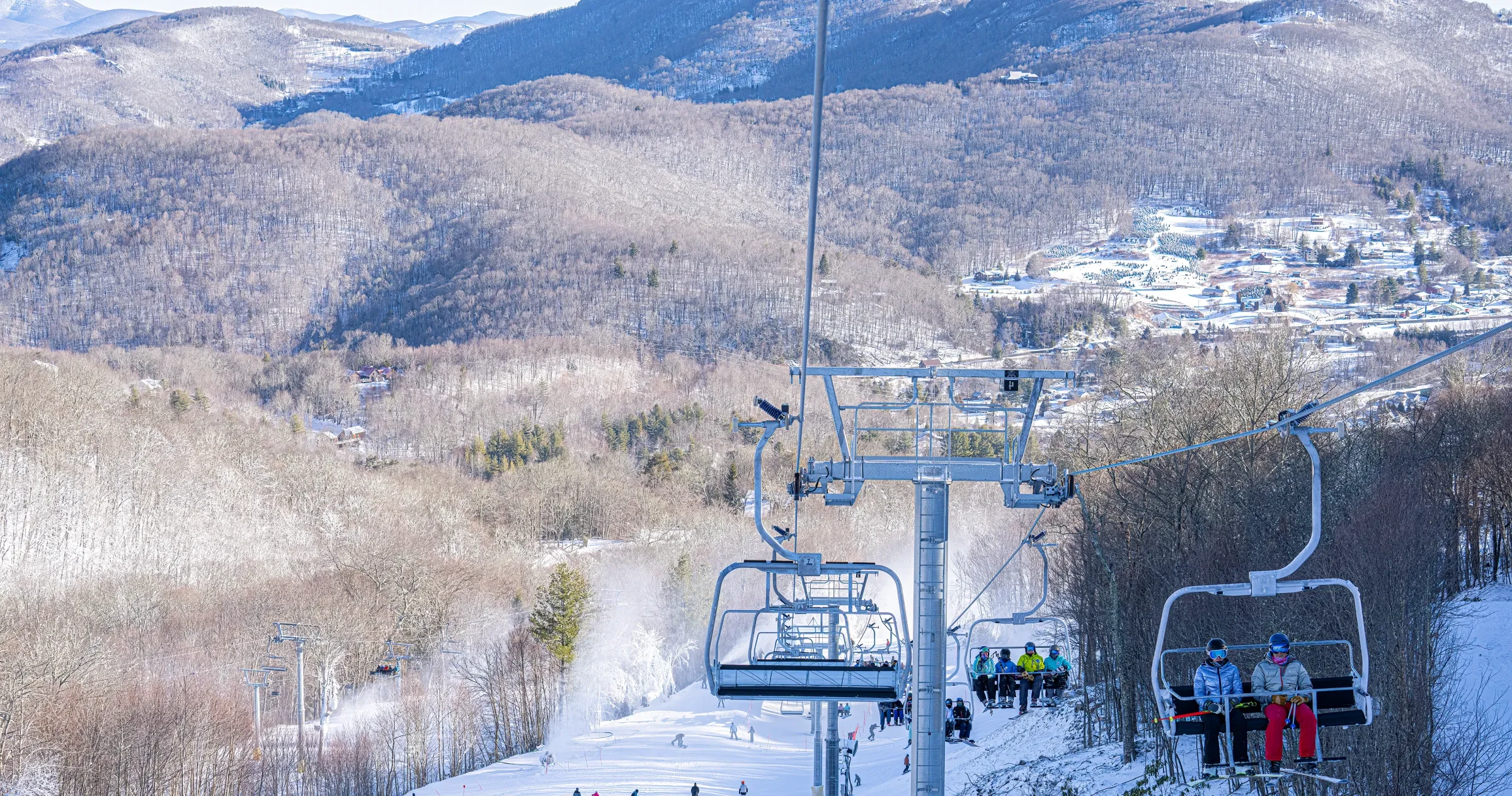 People on ski lift, skiiers on mountain below and big mountain in background.