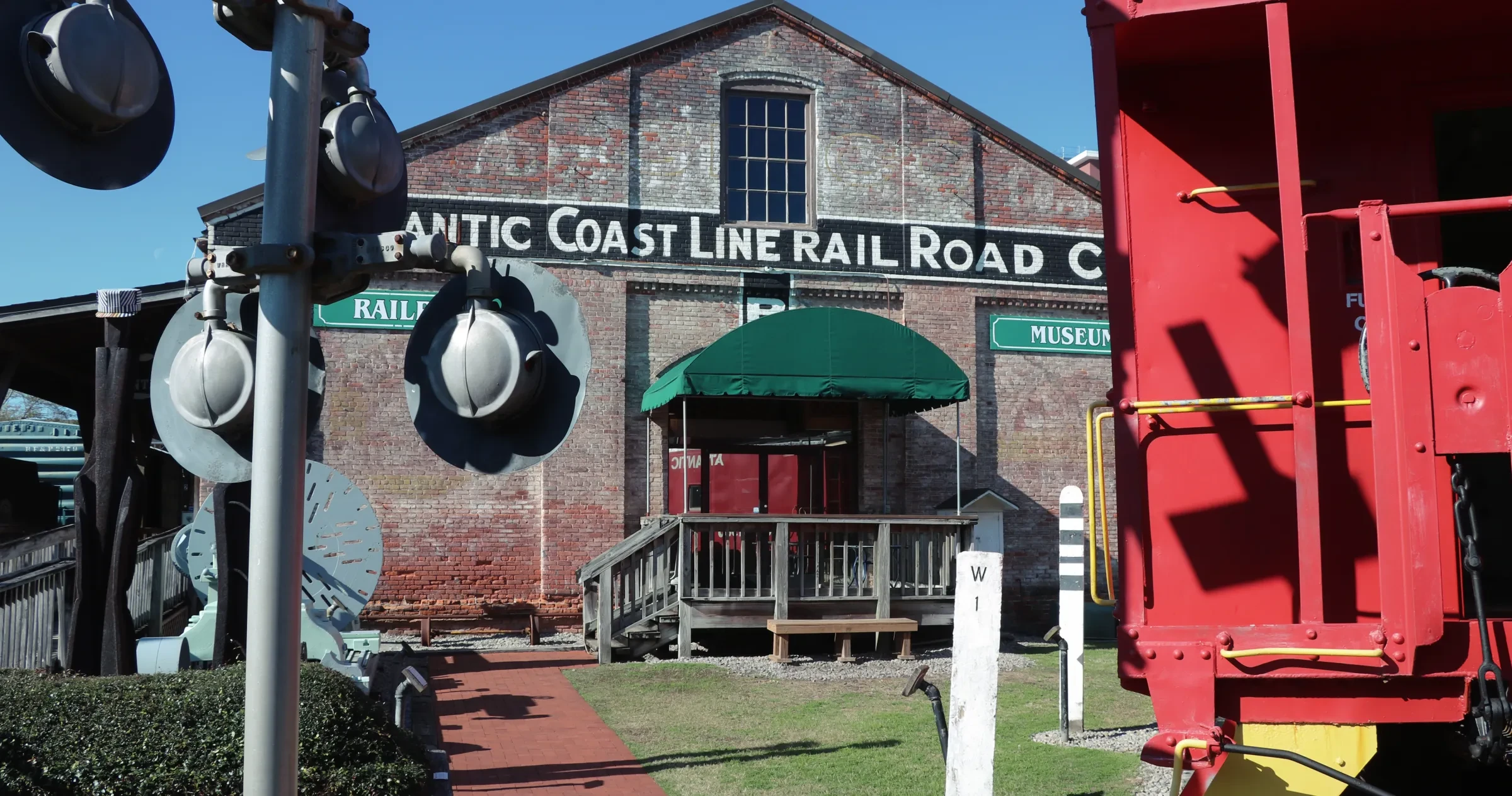 Old brick building with faded "Atlantic Coast Line Railroad" sign, green awning, and steps. Railroad crossing signals and a red train car are in the foreground, with clear blue sky above.