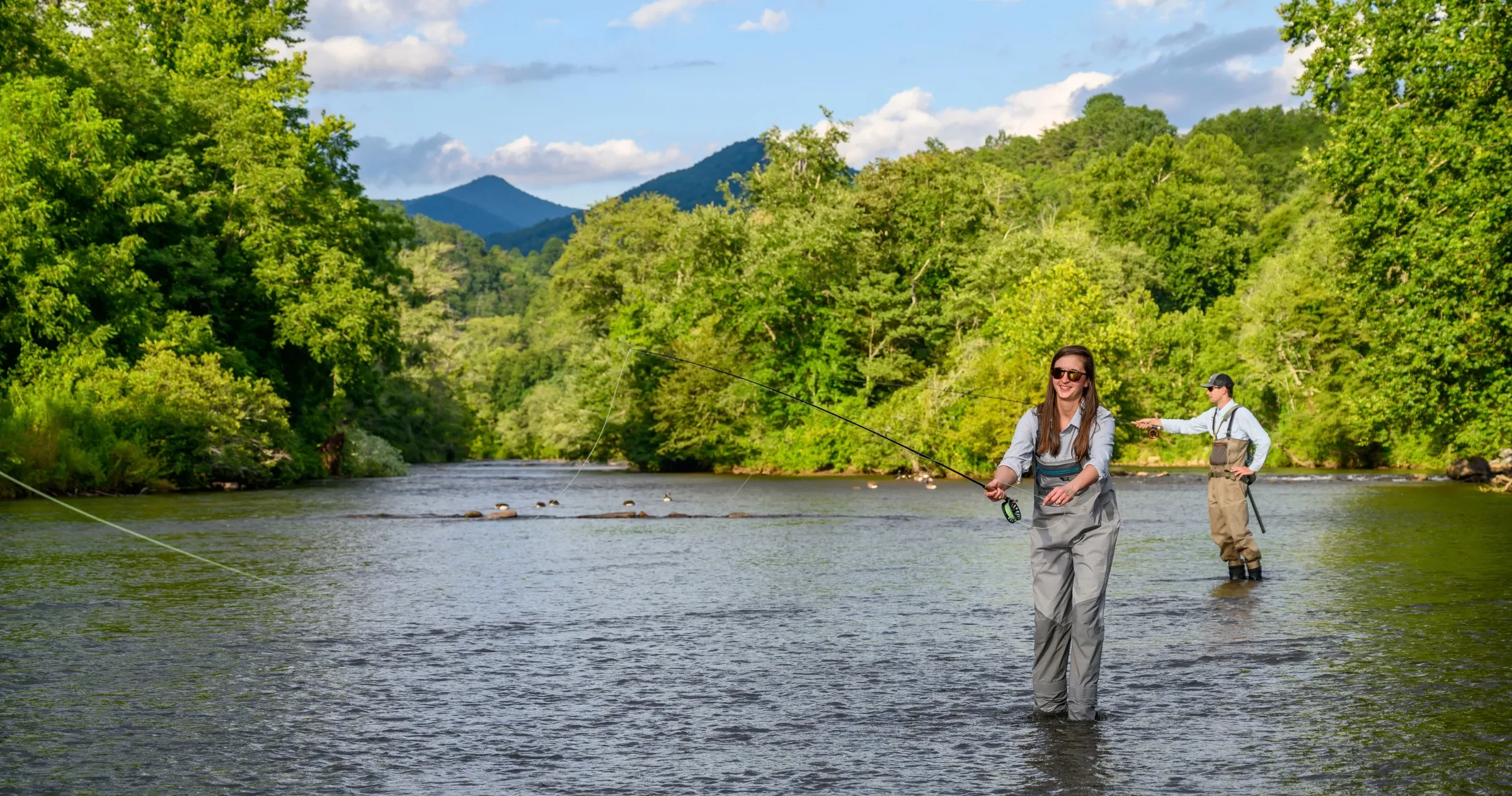 Two people fly-fishing in NC river with mountains in distance.
