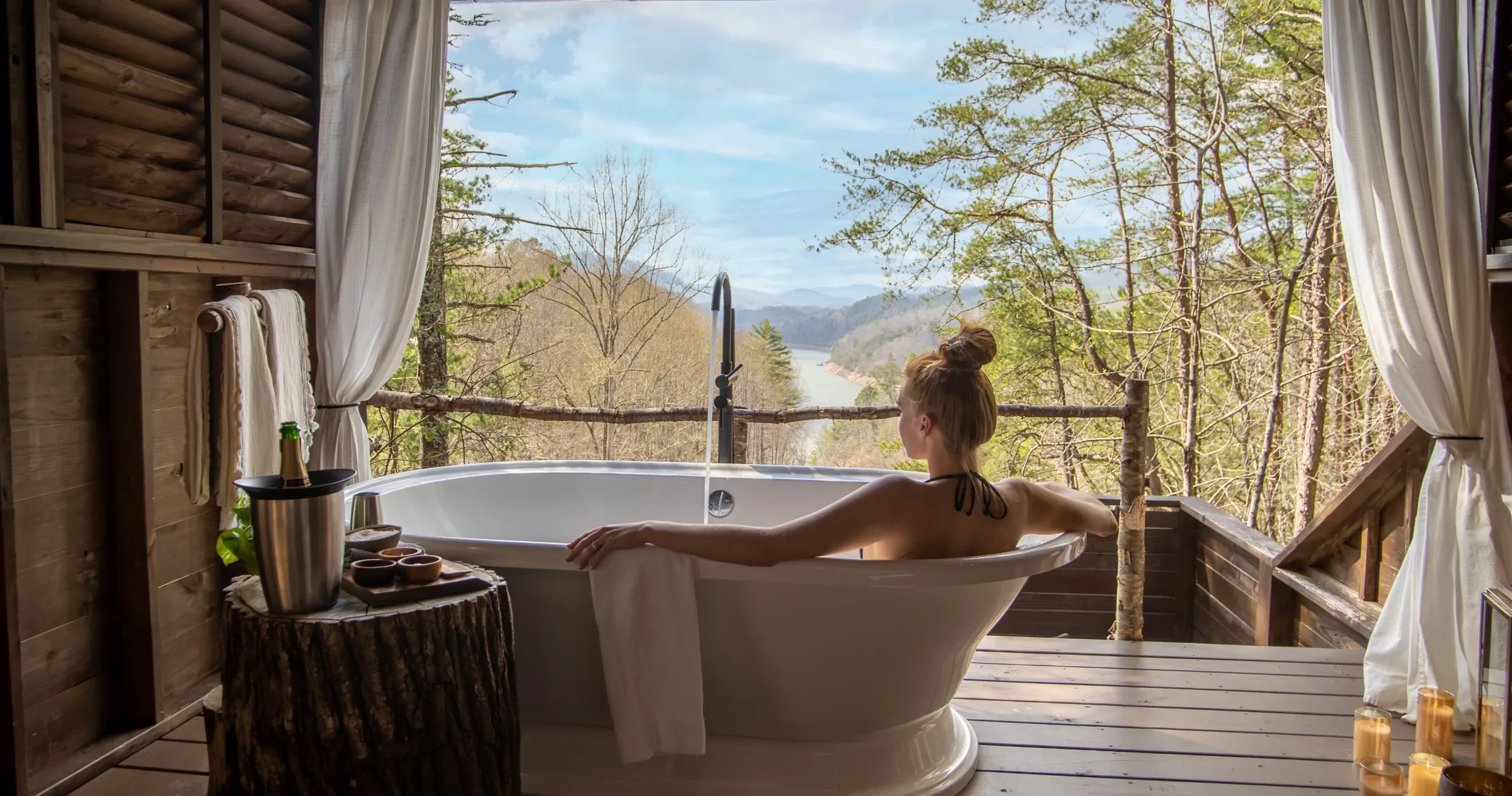 Woman in soaking tub in treehouse with forest, mountains and river in distance.