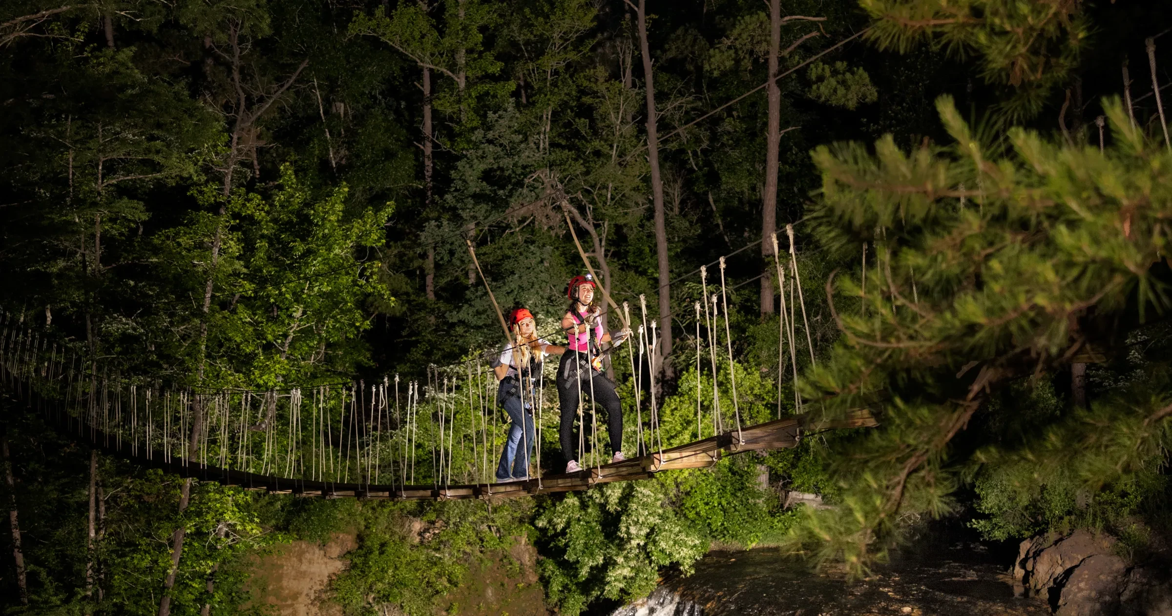 Two people on suspension bridge crossing over river at night on zip line tour.
