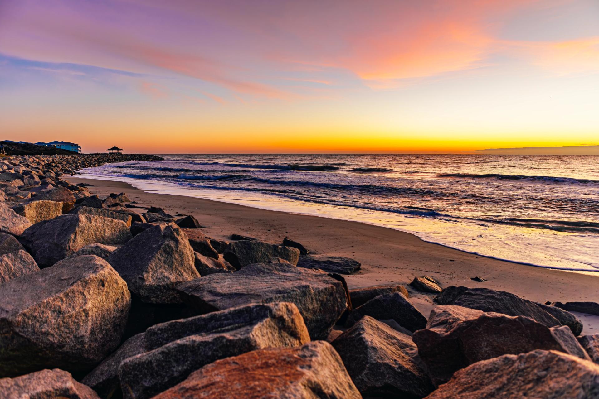 Sun rising over ocean on horizon with large rocks and sand in foreground.