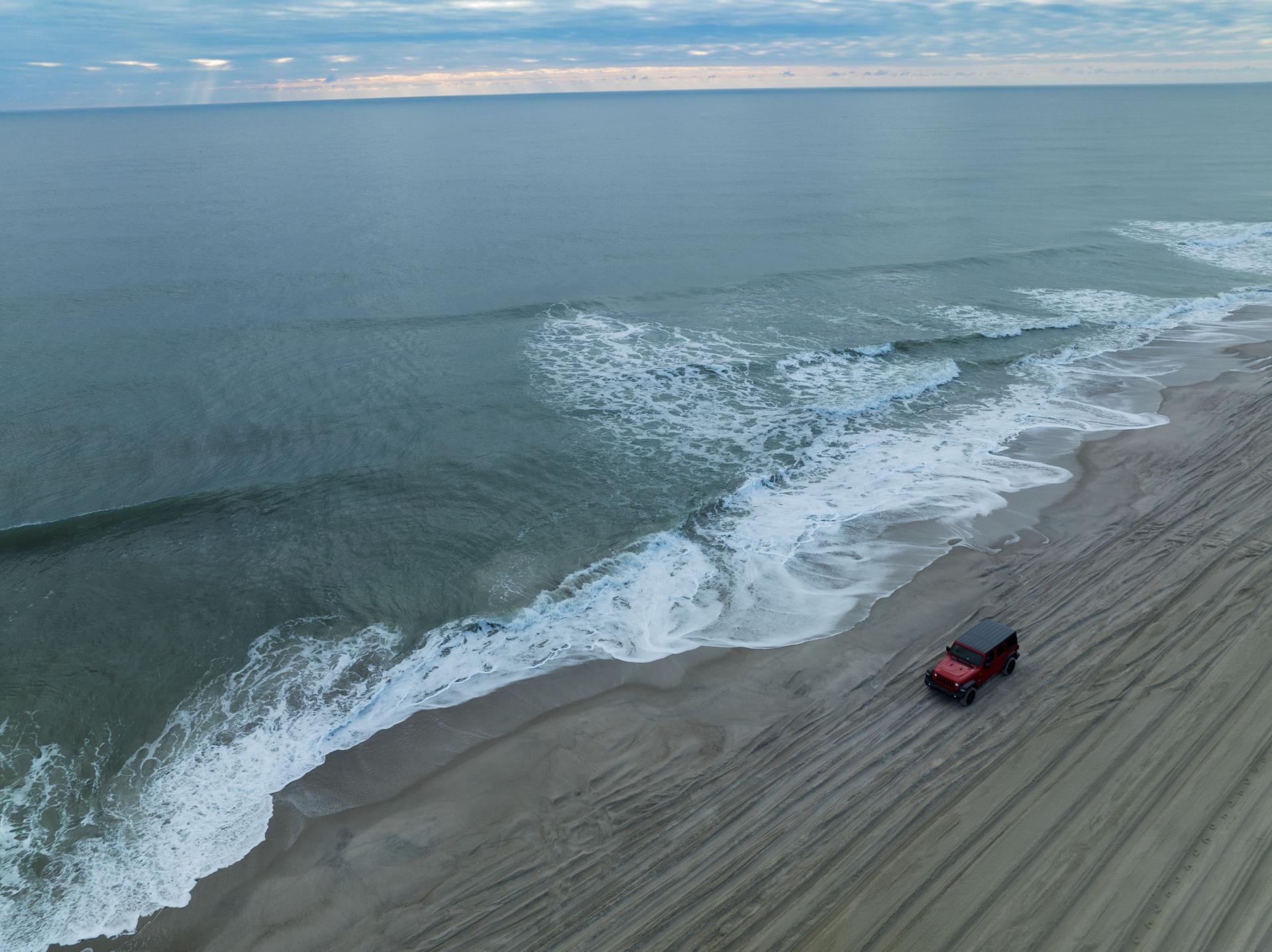Drone shot of jeep driving on beach with ocean to the left in winter.