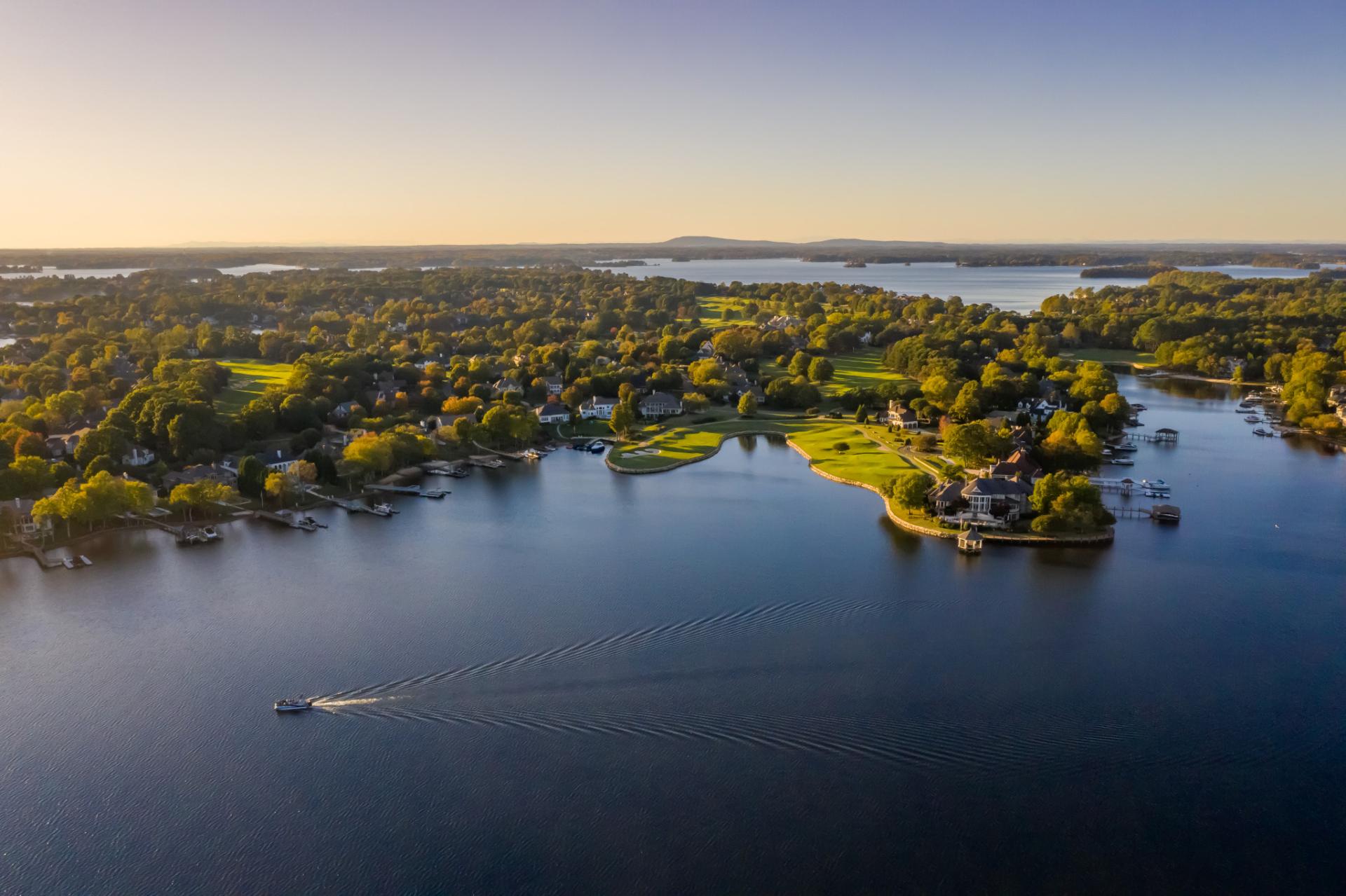 A lone boat motors across Lake Norman during golden hour. The shore is lined with green trees and the sky is an ombre color from yellow to deep blue.