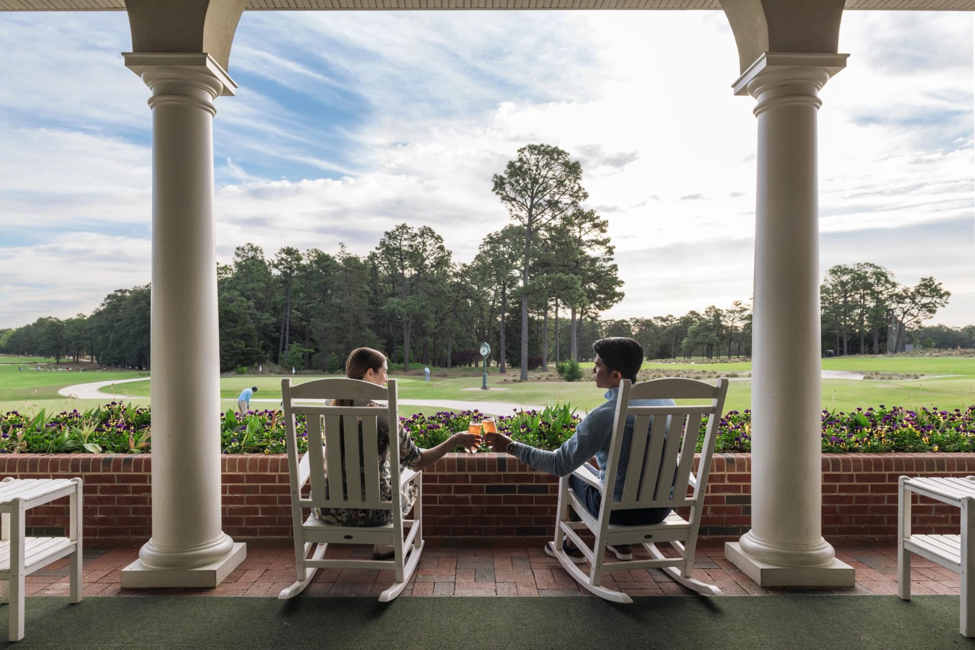 Back of couple sitting in rocking chairs on Pinehurst Resort porch, cheersing a cocktail, with golf grounds in background.