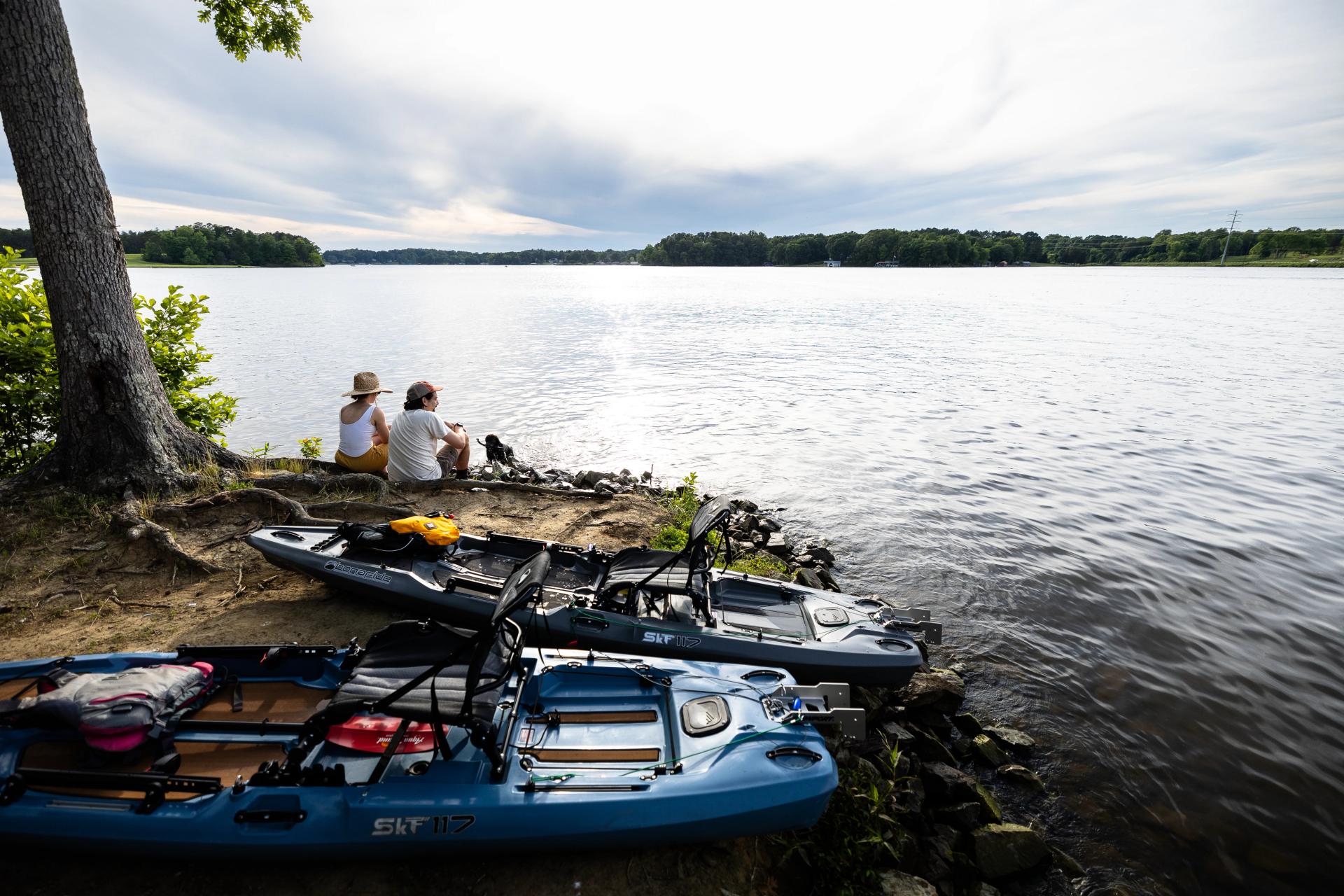 Couple sitting on edge of Hyco Lake admiring the view with kayaks in foreground on land.
