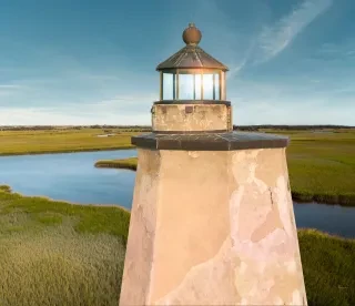 Aerial closeup view of Old Baldy Lighthouse exterior on Bald Head Island during daytime under wispy cloudy sky with view of sound and marsh below.