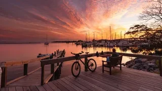tMan sitting on deck watching sunset over Oriental with his bike leaning against railing.