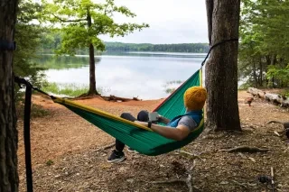 A camper lounges in a colorful hammock, sipping from a mug while gazing out at the calm lake. Tall trees frame the peaceful shoreline, with fishing gear and a campfire setup nearby.