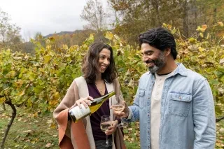Woman smiling while pouring white wine into her partner's glass within row of grapes at Grandfather Vineyard & Winery.