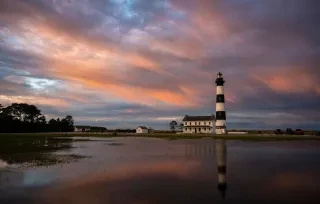 Bodie Island Lighthouse and house just right from center frame, with reflection of lighthouse and sunset clouds in the water.