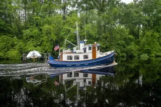 Horizontal shot of boat moving through calm Dismal Swamp Canal with green trees and foliage in background during cloudy daytime.