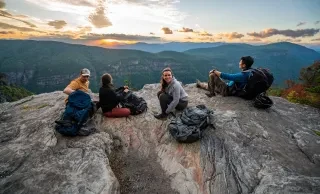 Group of four friend sitting on rock overlooking gorge and mountains with sunset in background.