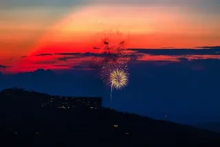 Fireworks show off against a vibrantly colored sunset with the shadow of mountains in the foreground.