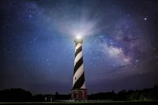 A dramatic image of Cape Hatteras Lighthouse lit up at night under a starry sky. 