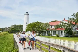 Side view of two women walking along pier near Ocracoke Island Lighthouse in daytime with building exterior in background.