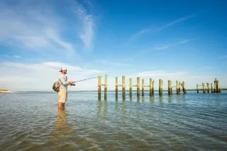 A man standing knee-deep in water, fishing near the remains of wooden pilings, with clear skies and calm surroundings.