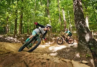 Two people on mountain bikes enjoy a thrilling turn at Ride Kanuga Mountain Biking Park amongst green forest.